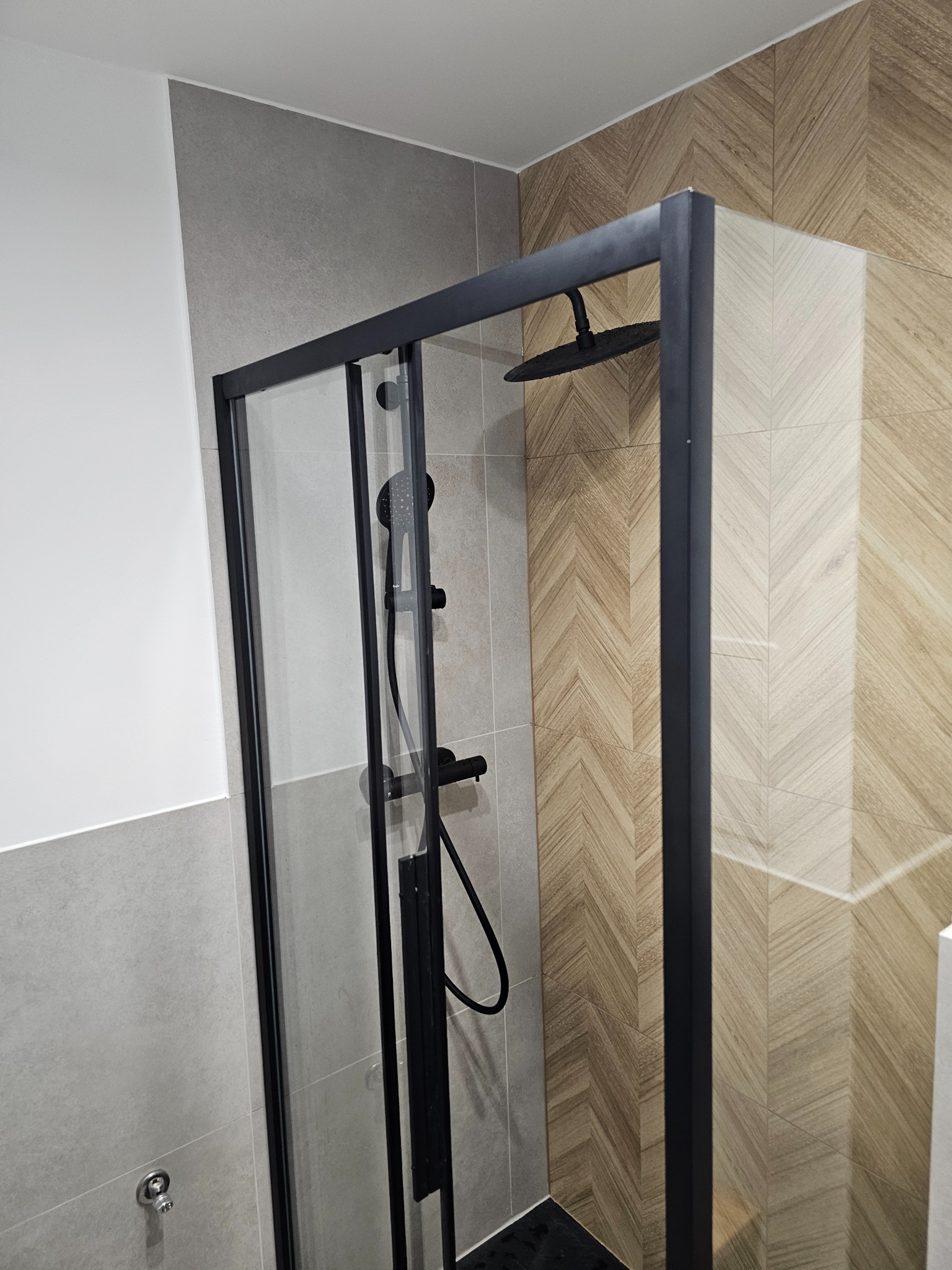 Nowoczesna kabina prysznicowa z czarną ramą, deszczownicą i baterią, wykończona płytkami w odcieniach szarości i drewna ułożonymi w jodełkę.