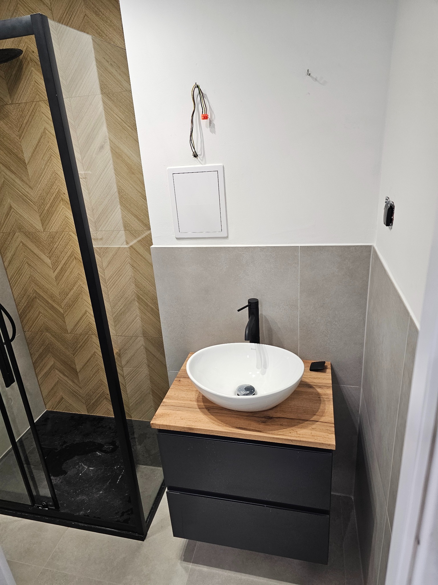 Nowoczesna łazienka z kabiną prysznicową typu walk-in z czarnymi profilami, umywalką nablatową na szafce z drewnianym blatem, szare płytki na ścianach i podłodze, widoczne niedokończone podłączenia...