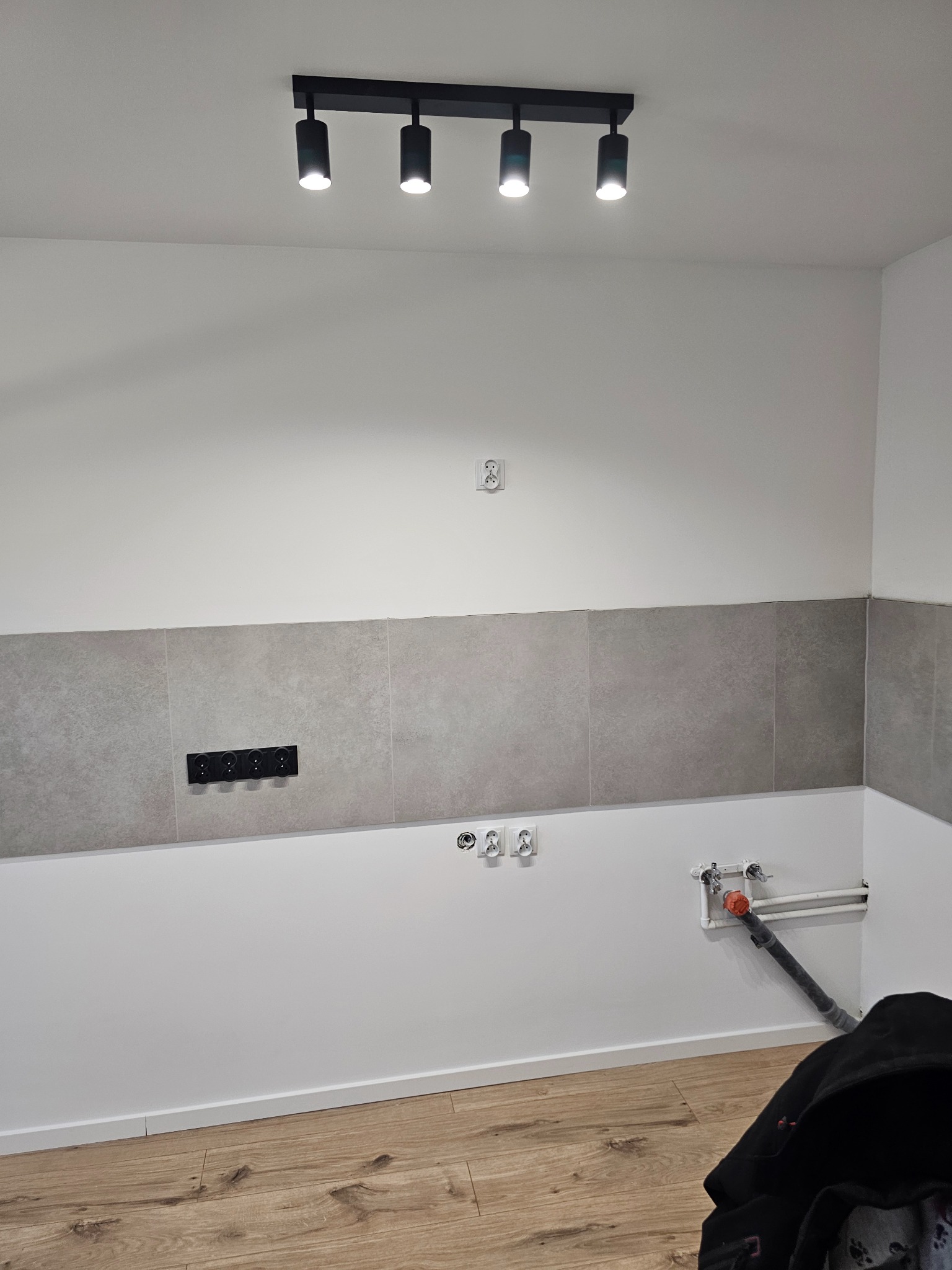 Ściana w trakcie wykańczania z szarymi płytkami, widoczne gniazdka elektryczne i instalacja wodno-kanalizacyjna, oświetlenie sufitowe punktowe.