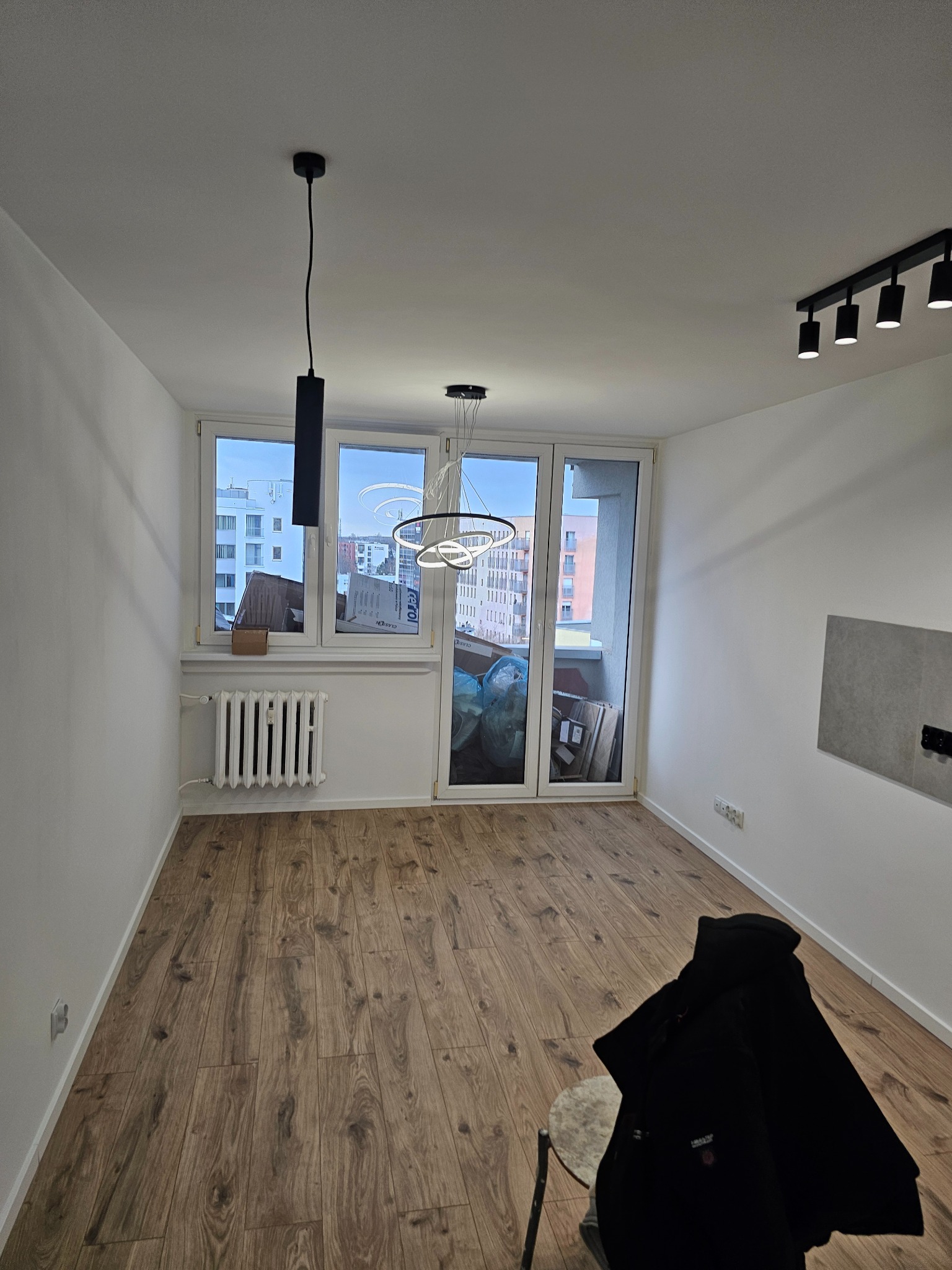 Pusty pokój z drewnianą podłogą, białymi ścianami, grzejnikiem pod oknem balkonowym, nowoczesnym oświetleniem sufitowym i czarną kurtką na krześle.