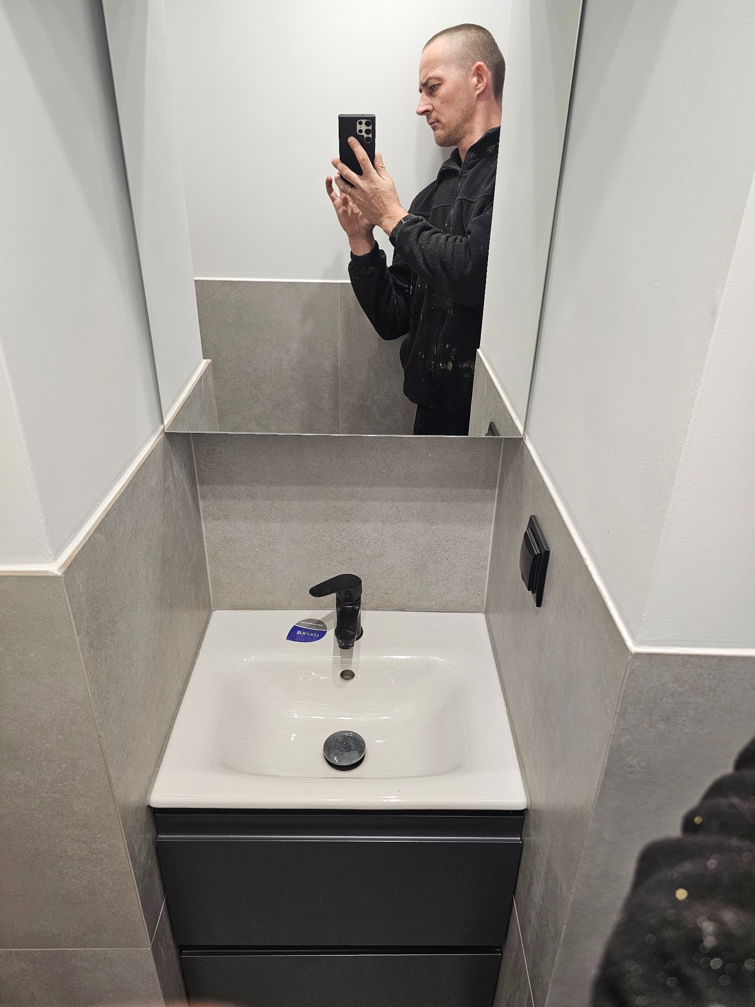 Nowoczesna łazienka po remoncie: biała umywalka z czarną armaturą i szafką w kolorze antracytu, szare płytki na ścianach, odbicie fotografa w lustrze.