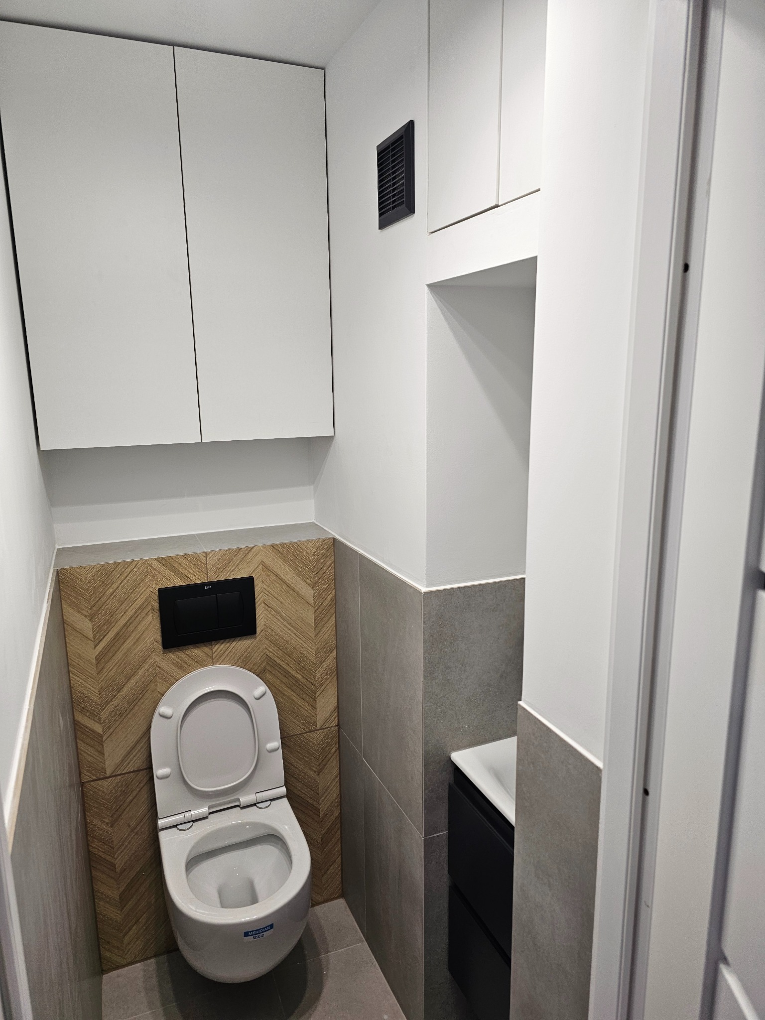 Nowoczesna, minimalistyczna toaleta z podwieszaną muszlą, drewnopodobną mozaiką ułożoną w jodełkę nad spłuczką i szarą płytką, białe szafki wiszące i czarna umywalka w tle.