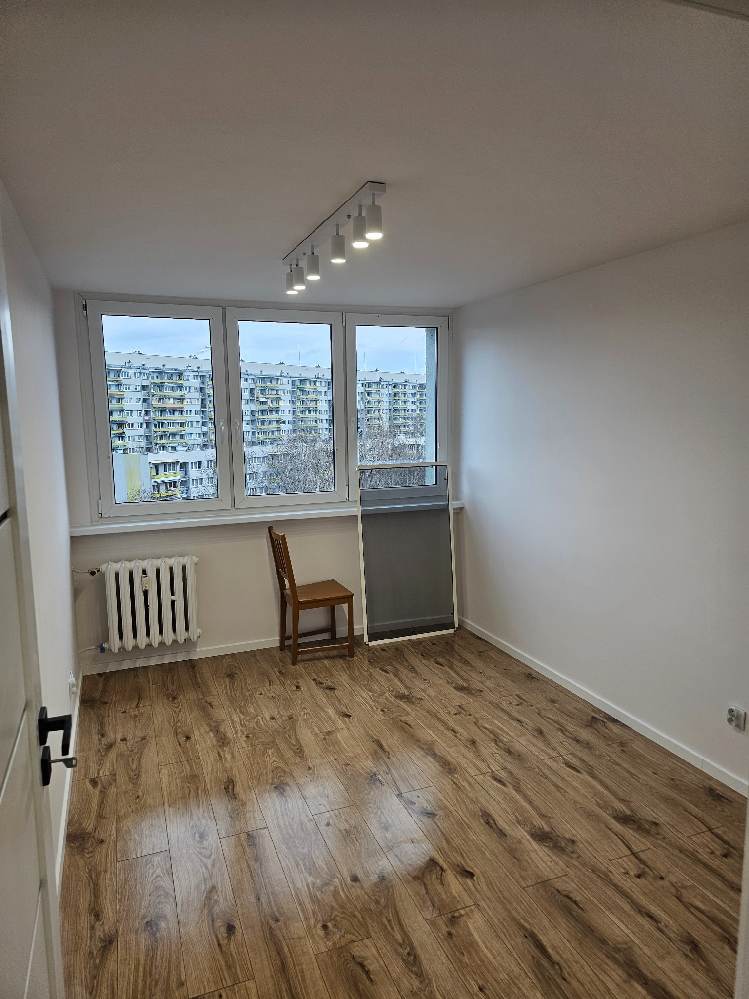 Pusty pokój po remoncie z drewnianą podłogą, białymi ścianami i oknem z widokiem na blokowisko, uzupełniony krzesłem i grzejnikiem.