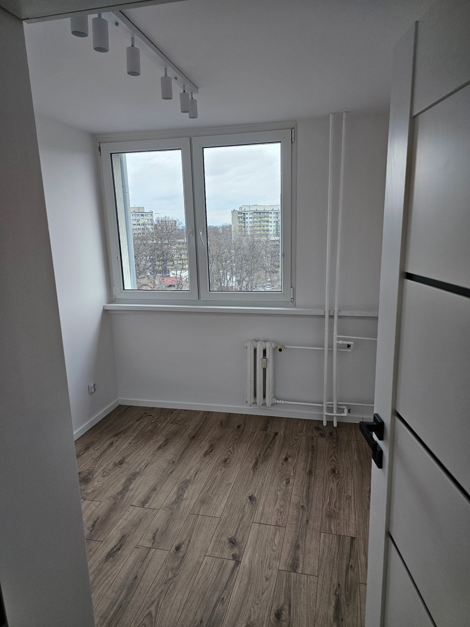 Jasne wnętrze pokoju po remoncie z drewnopodobną podłogą, białymi ścianami, grzejnikiem i widokiem na blok z okna.