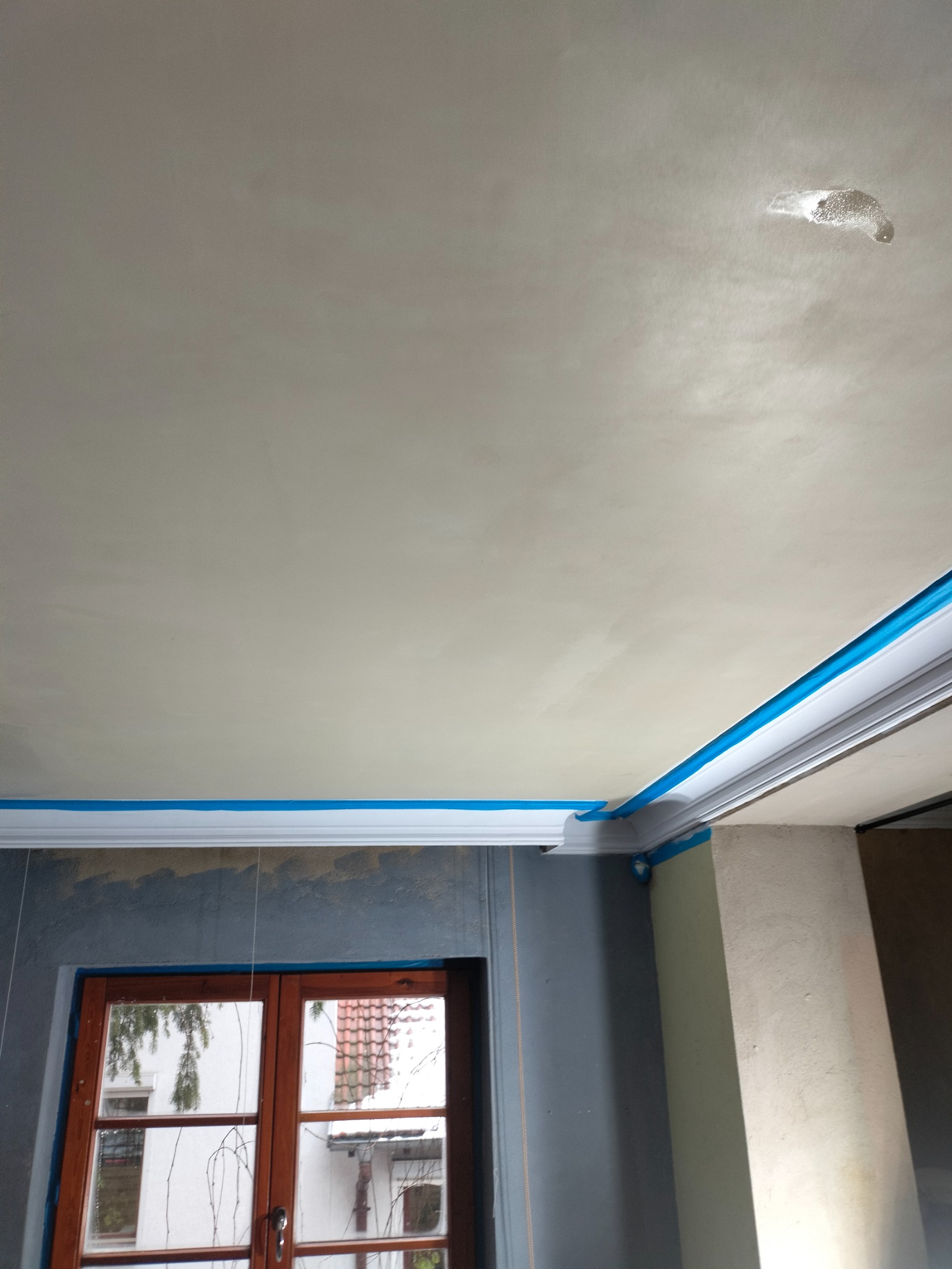 Świeżo położona, jasna gładź na suficie z widocznym, delikatnym cieniowaniem, zabezpieczona niebieską taśmą malarską przy ozdobnej listwie sufitowej i framudze okna.