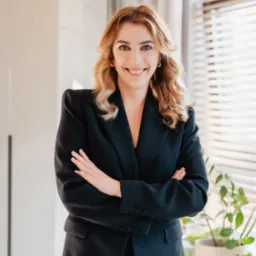 Uśmiechnięta kobieta w czarnym garniturze, z założonymi rękami, stojąca w jasnym biurze obok biurka z książkami i rośliną doniczkową przy oknie z żaluzjami.