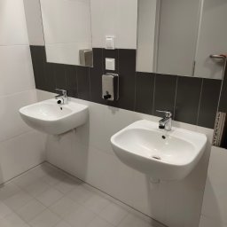 AW SANITARNE SP Z O.O. - Dwie białe umywalki zamontowane na ścianie w łazience z szaro-białymi płytkami, widoczne baterie, dozownik mydła i fragment lustra.
