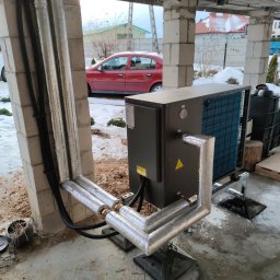 AW SANITARNE SP Z O.O. - Szara pompa ciepła zamontowana na zewnątrz budynku w trakcie instalacji, rury owinięte folią izolacyjną, widoczne elementy konstrukcyjne budynku i czerwony samochód w tle.