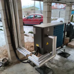 Szara pompa ciepła zamontowana na zewnątrz budynku w trakcie instalacji, rury owinięte folią izolacyjną, widoczne elementy konstrukcyjne budynku i czerwony samoch&oacute;d w tle.