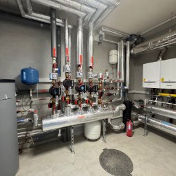AW SANITARNE SP Z O.O. - Węzeł cieplny w kotłowni: liczne rury w srebrnej izolacji, pompy obiegowe Grundfos, manometry, zbiorniki wyrównawcze, grzejniki i gaśnica w tle.