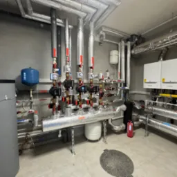 Węzeł cieplny w kotłowni: liczne rury w srebrnej izolacji, pompy obiegowe Grundfos, manometry, zbiorniki wyrównawcze, grzejniki i gaśnica w tle.