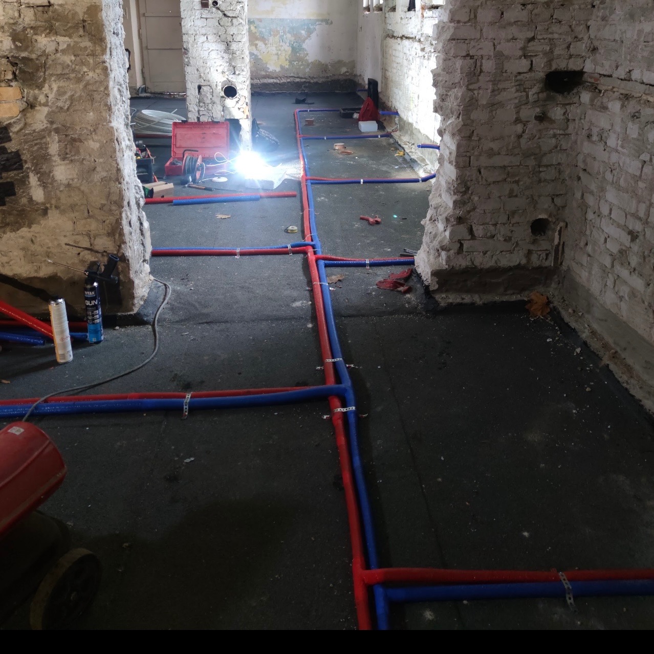Instalacja rur wodnych w surowym wnętrzu, widoczne czerwone i niebieskie rury łączone złączkami, narzędzia pracy i spawarka w tle, ceglane ściany w stanie deweloperskim.