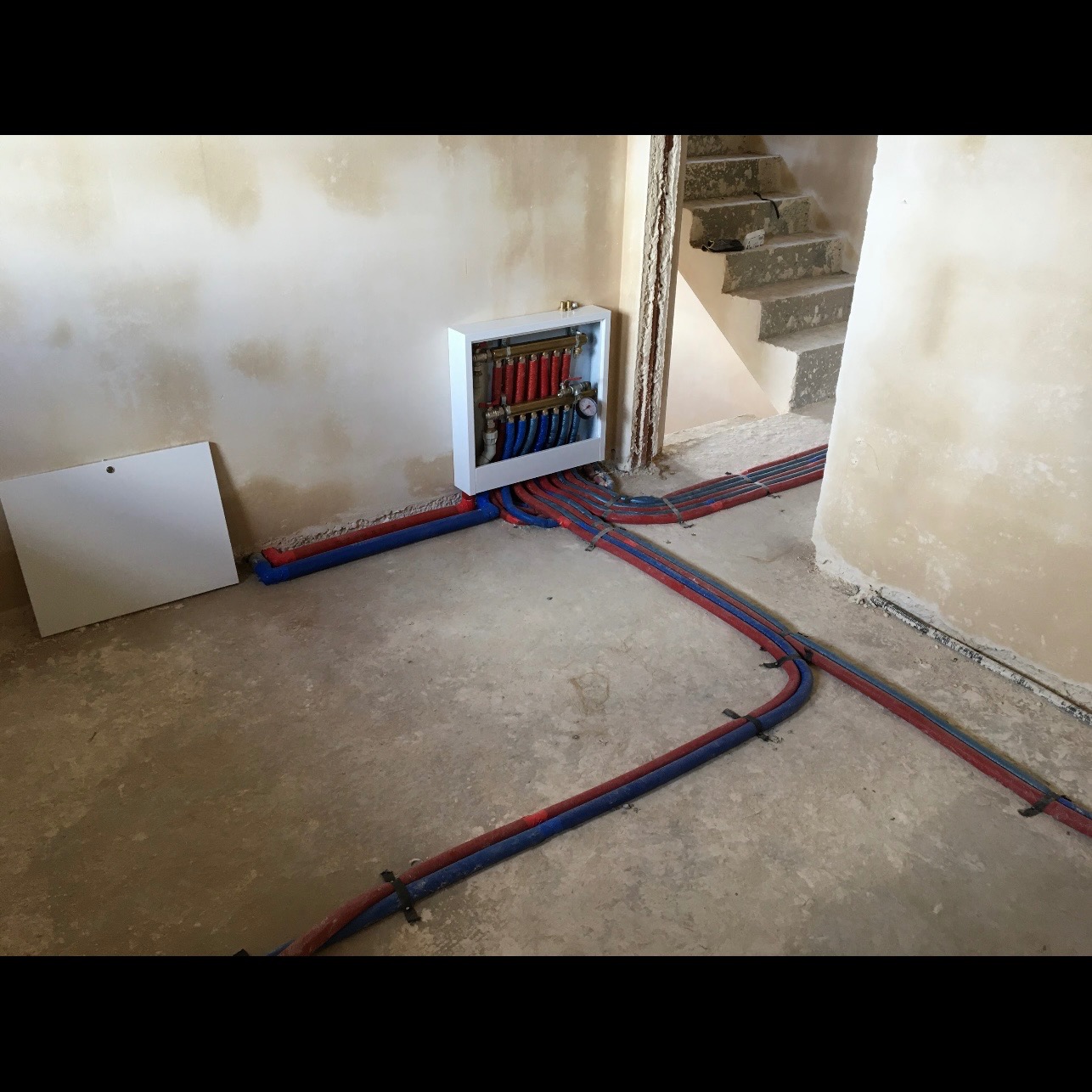 Rozdzielacz instalacji centralnego ogrzewania w białej obudowie, z licznymi rurami w kolorze czerwonym i niebieskim, ułożonymi na surowej podłodze w trakcie prac wykończeniowych, widoczne schody...