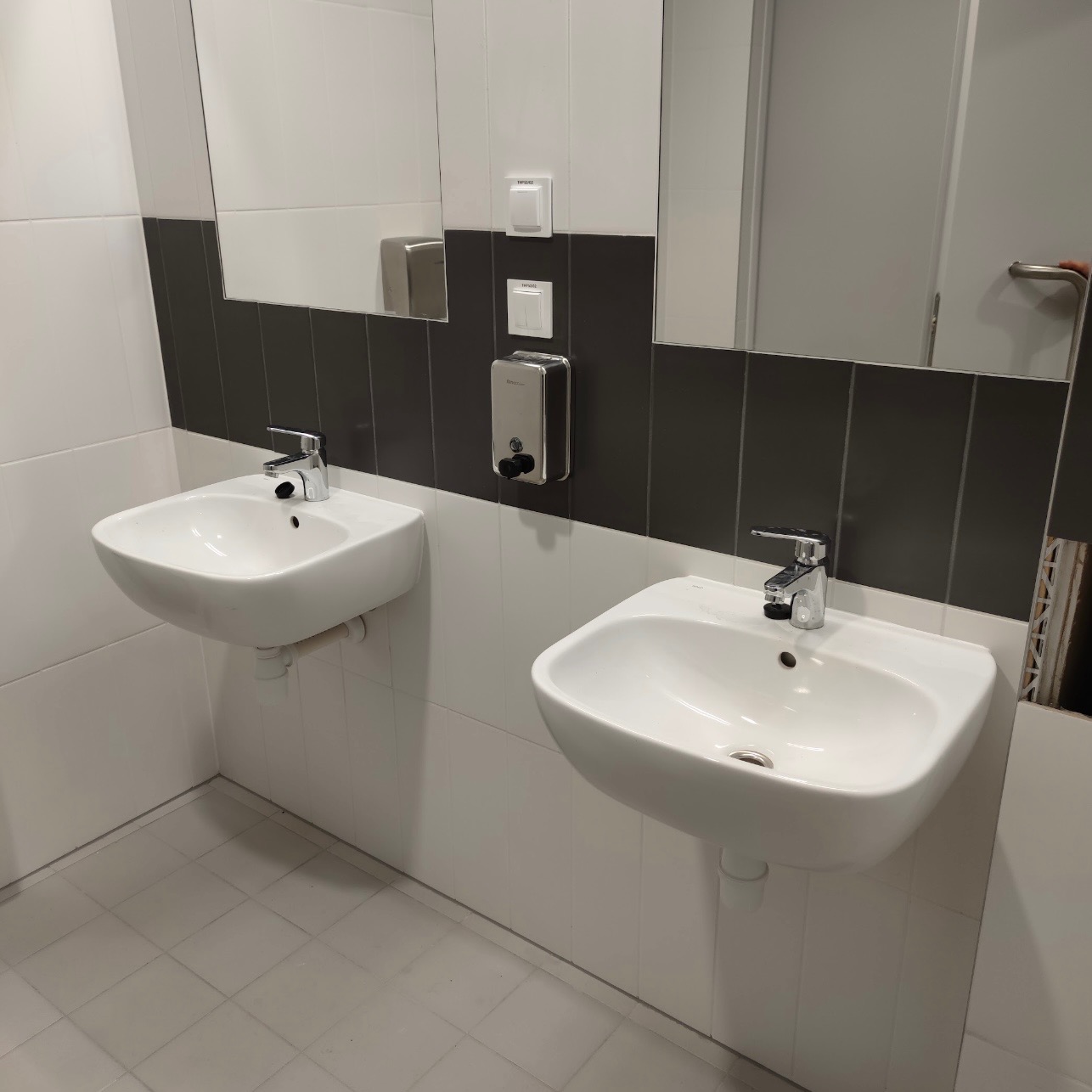 Dwie białe umywalki zamontowane na ścianie w łazience z szaro-białymi płytkami, widoczne baterie, dozownik mydła i fragment lustra.