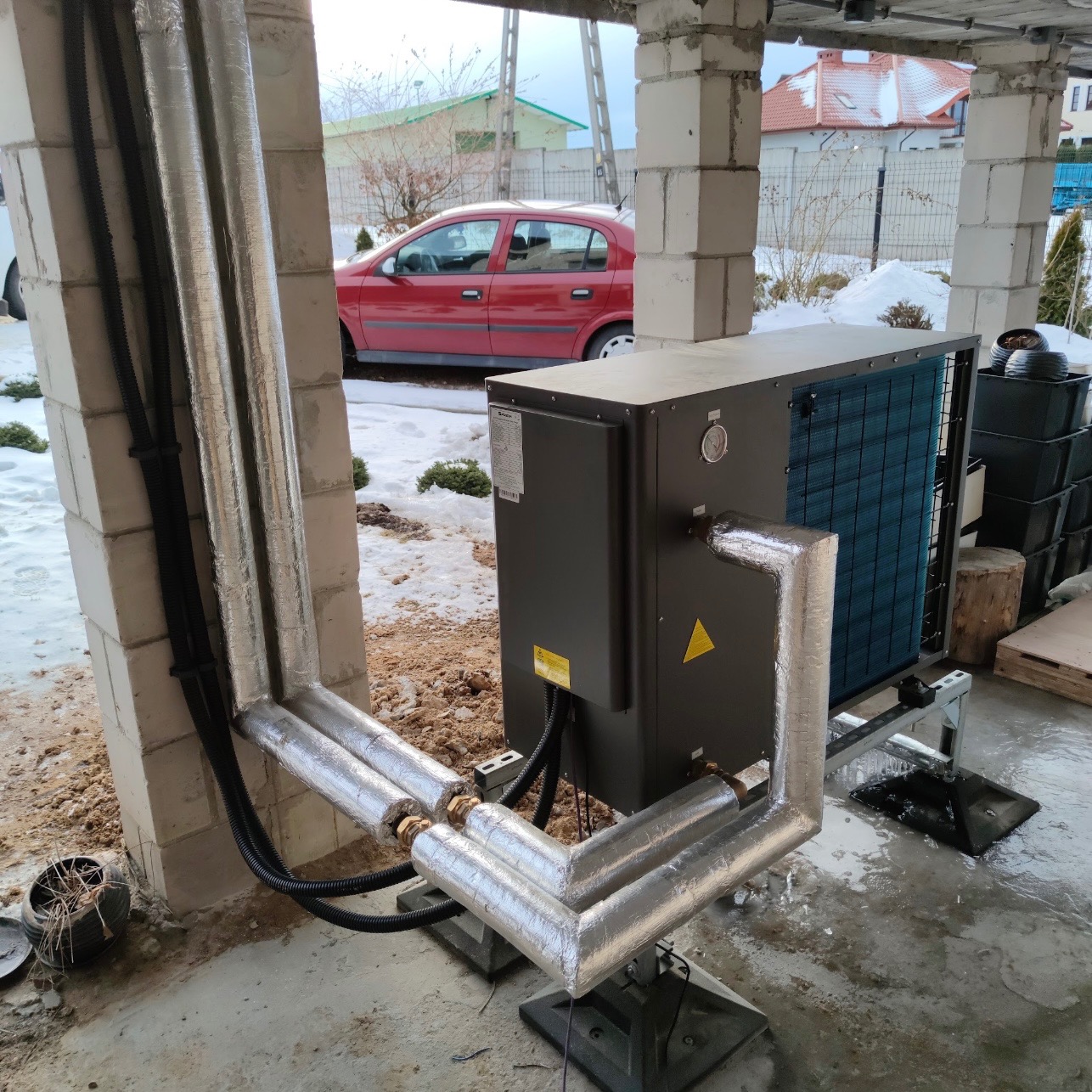 Szara pompa ciepła zamontowana na zewnątrz budynku w trakcie instalacji, rury owinięte folią izolacyjną, widoczne elementy konstrukcyjne budynku i czerwony samochód w tle.