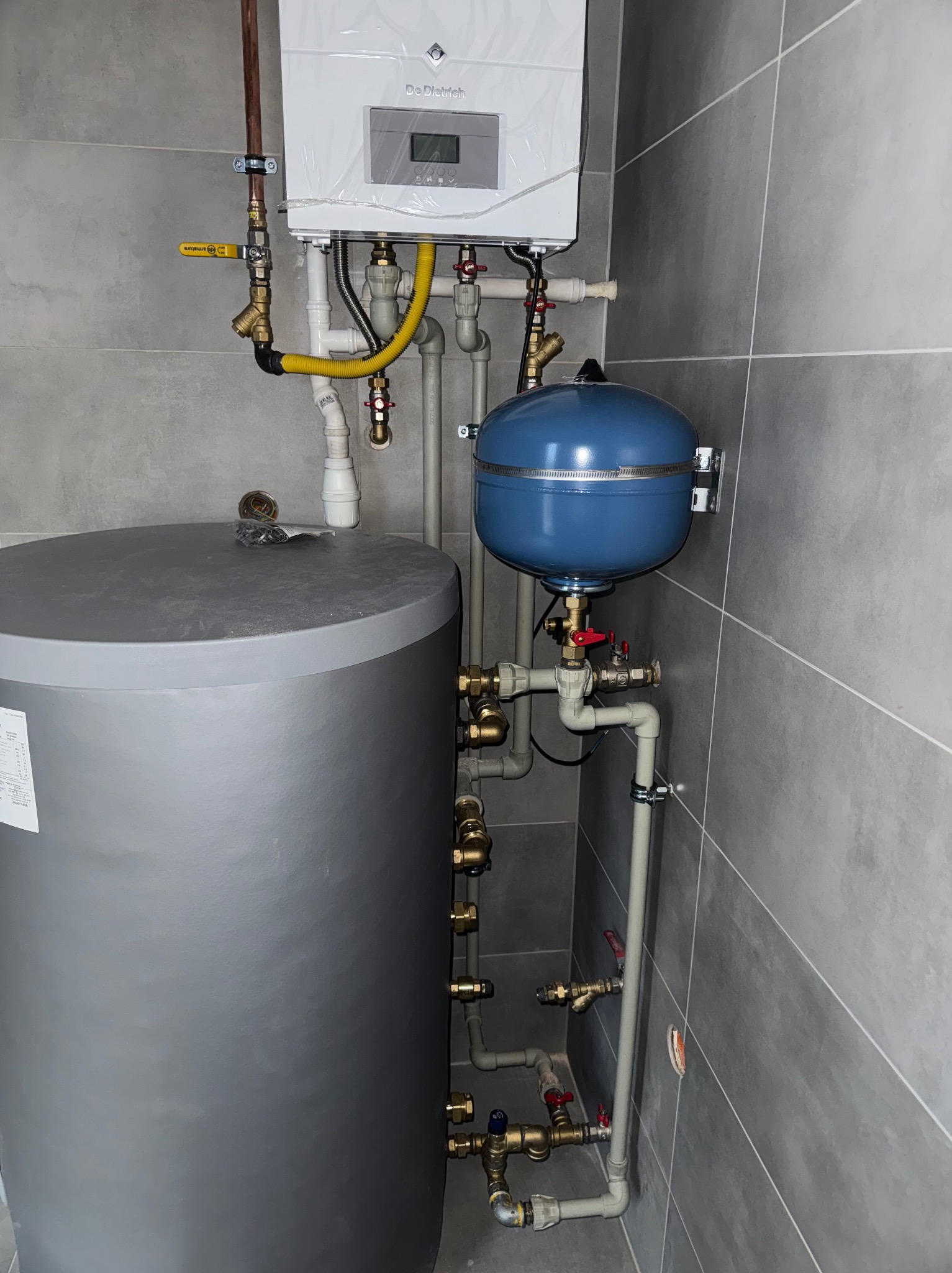 Nowy, biały piec gazowy marki De Dietrich z podłączonymi rurami miedzianymi i żółtym przewodem gazowym, obok szary zbiornik na wodę i niebieskie naczynie przeponowe w pomieszczeniu wyłożonym szarą...