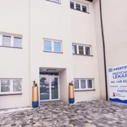 Fasada budynku przychodni lekarskiej Merkur Medica z bożonarodzeniową choinką i dekoracyjnymi donicami przed wejściem, widoczny szyld informacyjny z numerem telefonu.