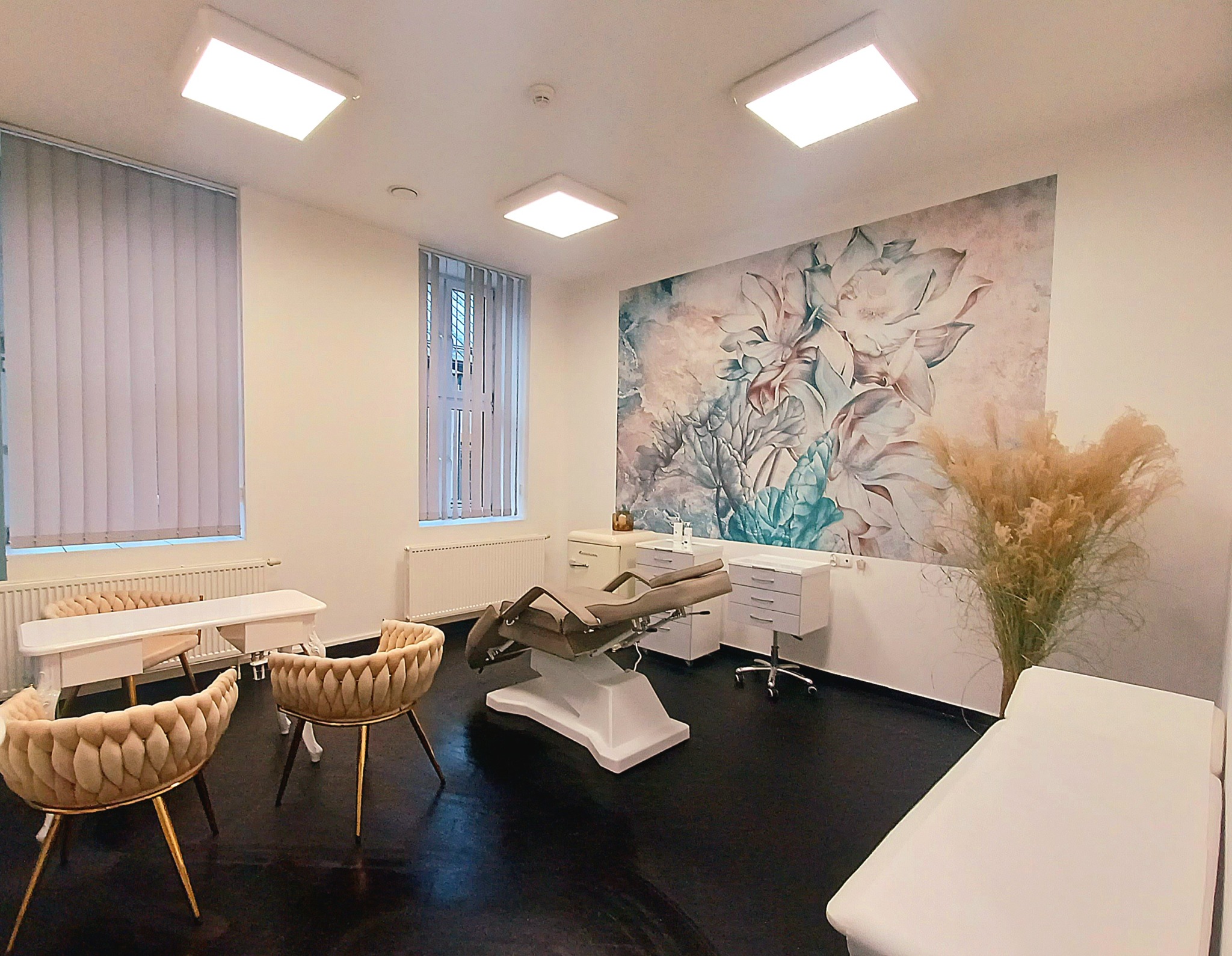 Jasne wnętrze gabinetu z fotelem zabiegowym, białymi meblami i dekoracyjną tapetą w kwiaty, oświetlone ledowymi panelami.