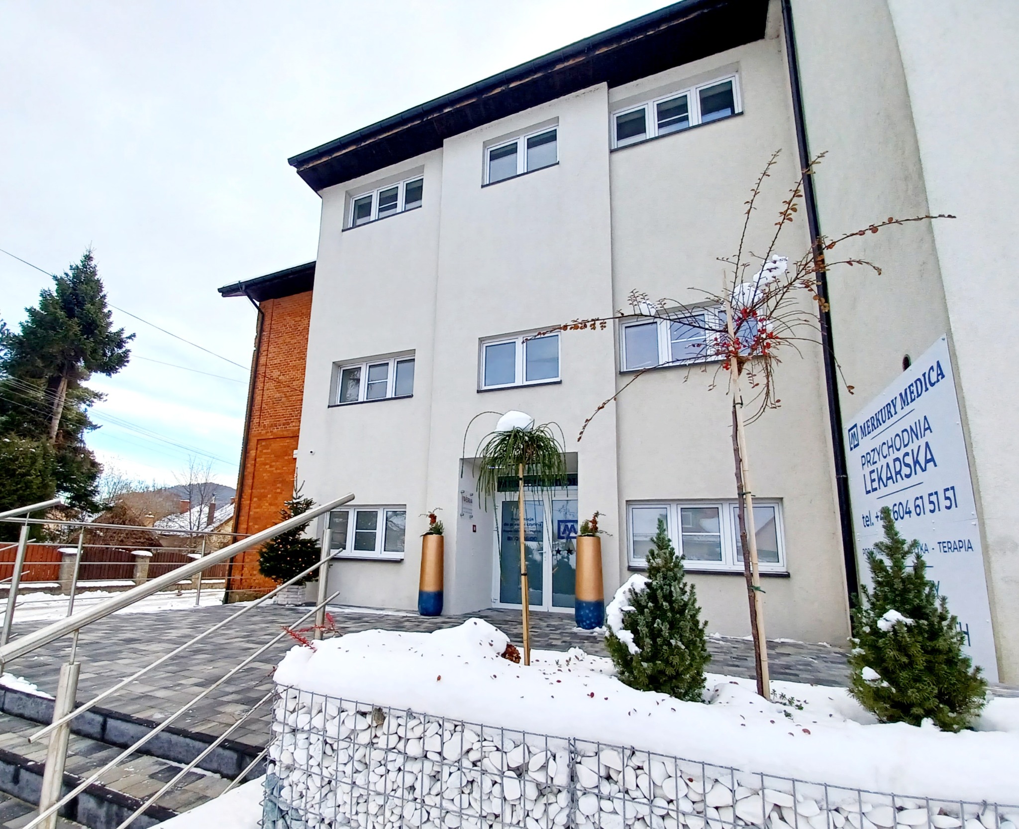 Budynek przychodni lekarskiej Merkury Medica w zimowej scenerii, widok z zewnątrz, z dekoracjami świątecznymi i śniegiem.