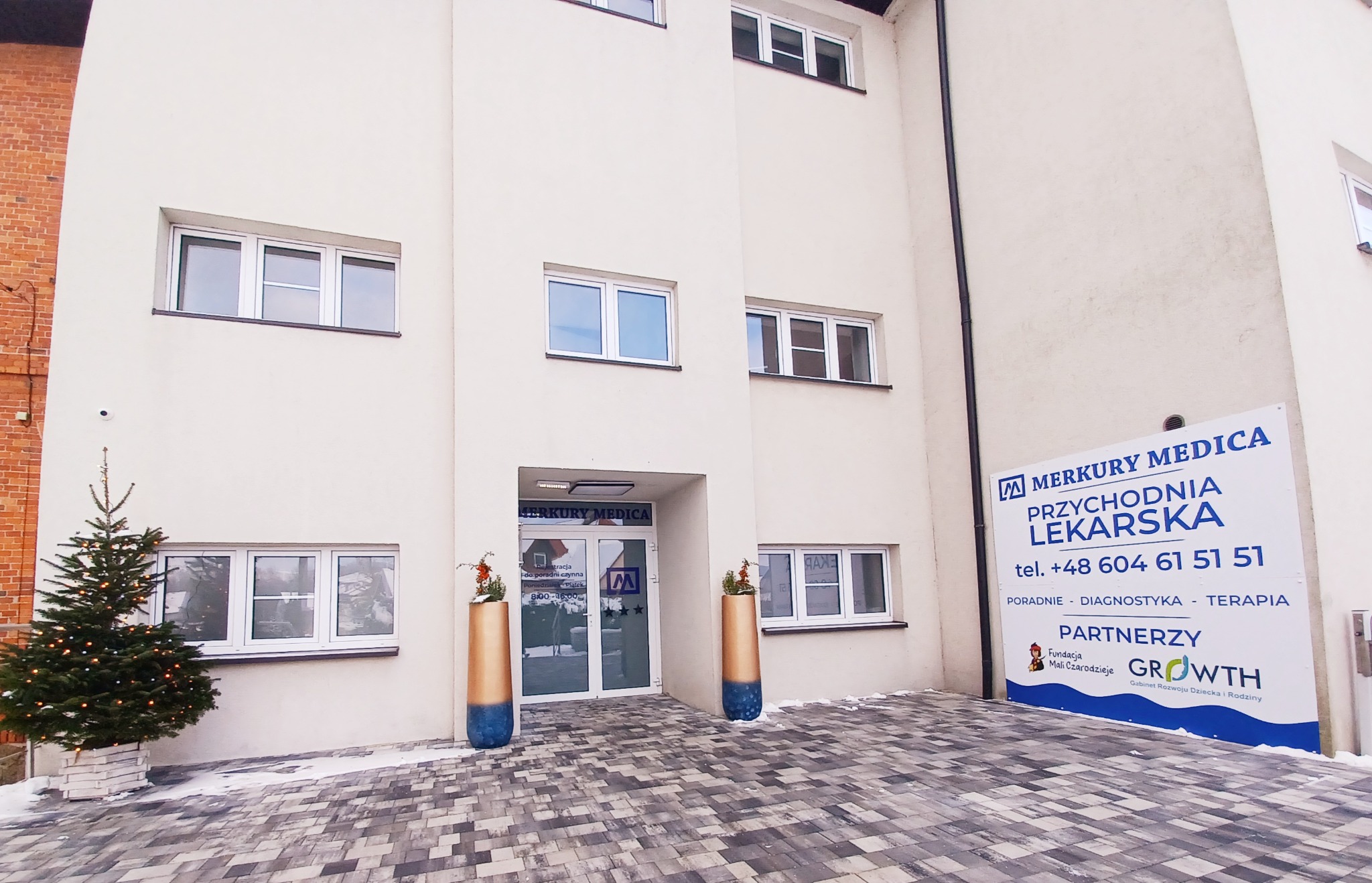 Fasada budynku przychodni lekarskiej Merkur Medica z bożonarodzeniową choinką i dekoracyjnymi donicami przed wejściem, widoczny szyld informacyjny z numerem telefonu.