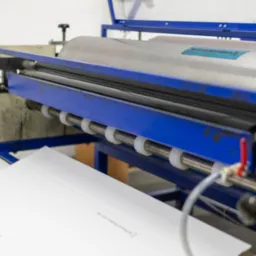 Maszyna drukarska z niebieską ramą aplikująca nadruk ze znakami ostrzegawczymi na białym opakowaniu kartonowym.