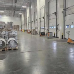 Wnętrze magazynu z paletami załadowanymi towarem w folii stretch, w tle widoczne bramy wjazdowe i w&oacute;zki widłowe.