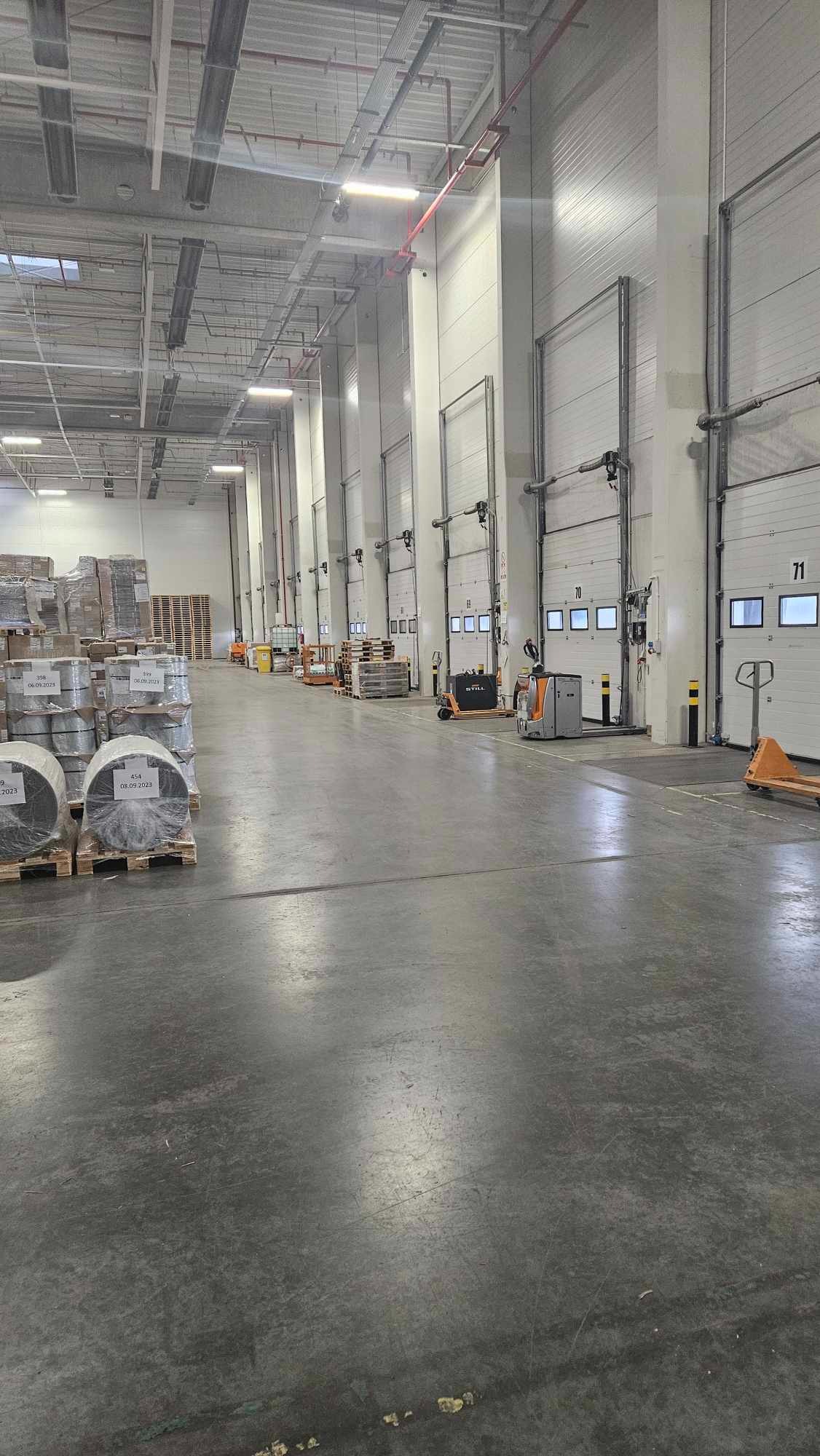 Wnętrze magazynu z paletami załadowanymi towarem w folii stretch, w tle widoczne bramy wjazdowe i wózki widłowe.