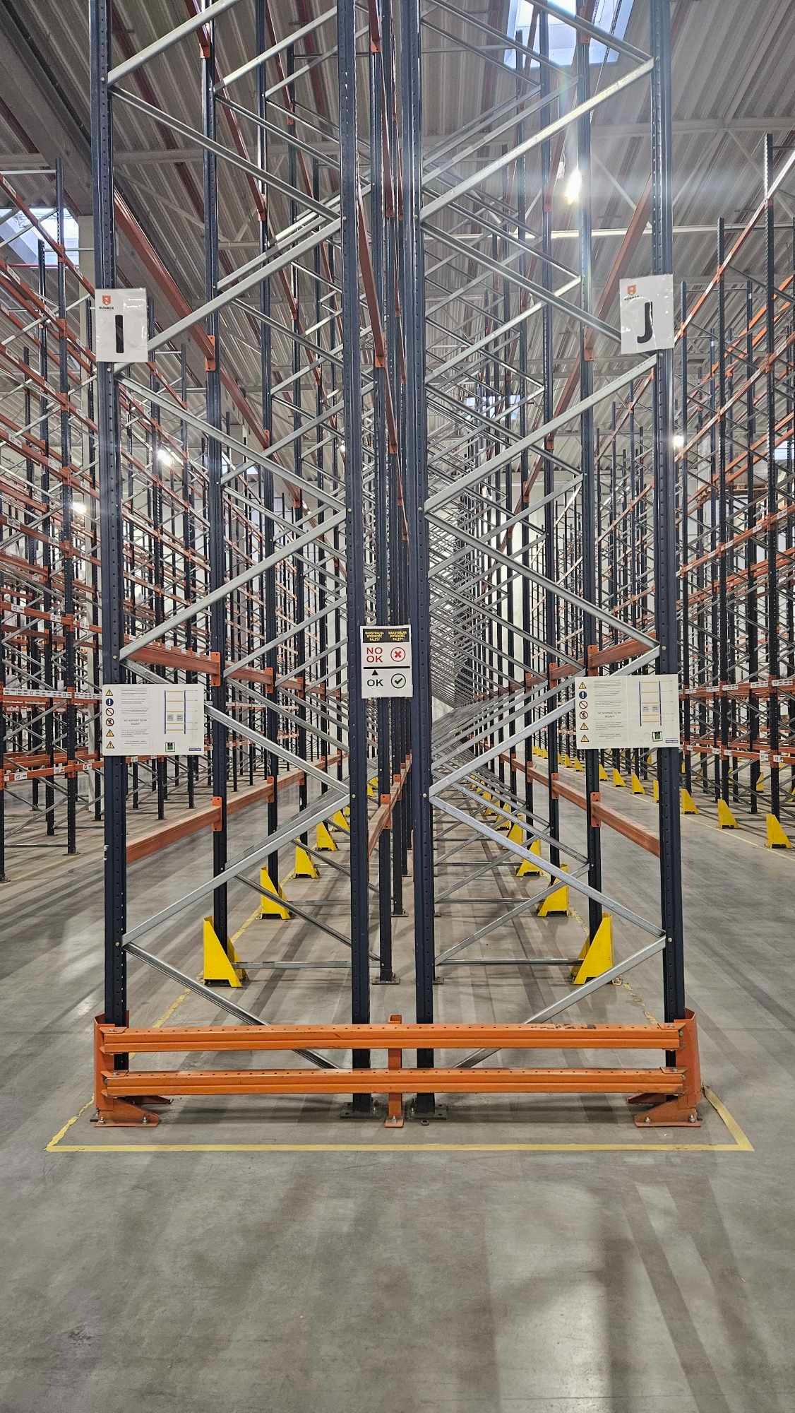 Puste regały magazynowe z czarnymi słupami i pomarańczowymi poprzeczkami, żółte osłony na nogach regałów, widoczne tabliczki informacyjne.