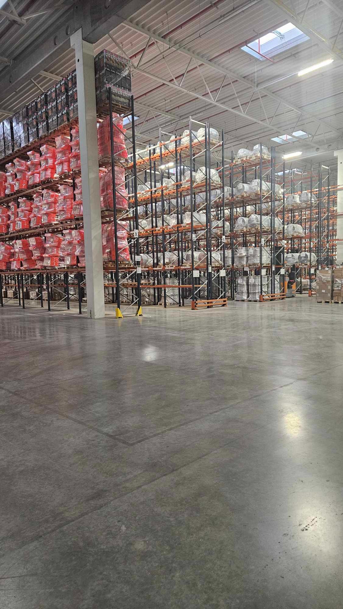 Wnętrze magazynu wysokiego składowania z regałami wypełnionymi towarem owiniętym folią, widok z poziomu posadzki.