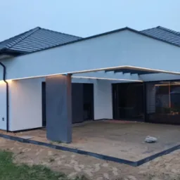 Nowoczesny dom jednorodzinny z oknami z żaluzjami zewnętrznymi, podświetlonym zadaszeniem tarasu i zagospodarowanym terenem wokół.
