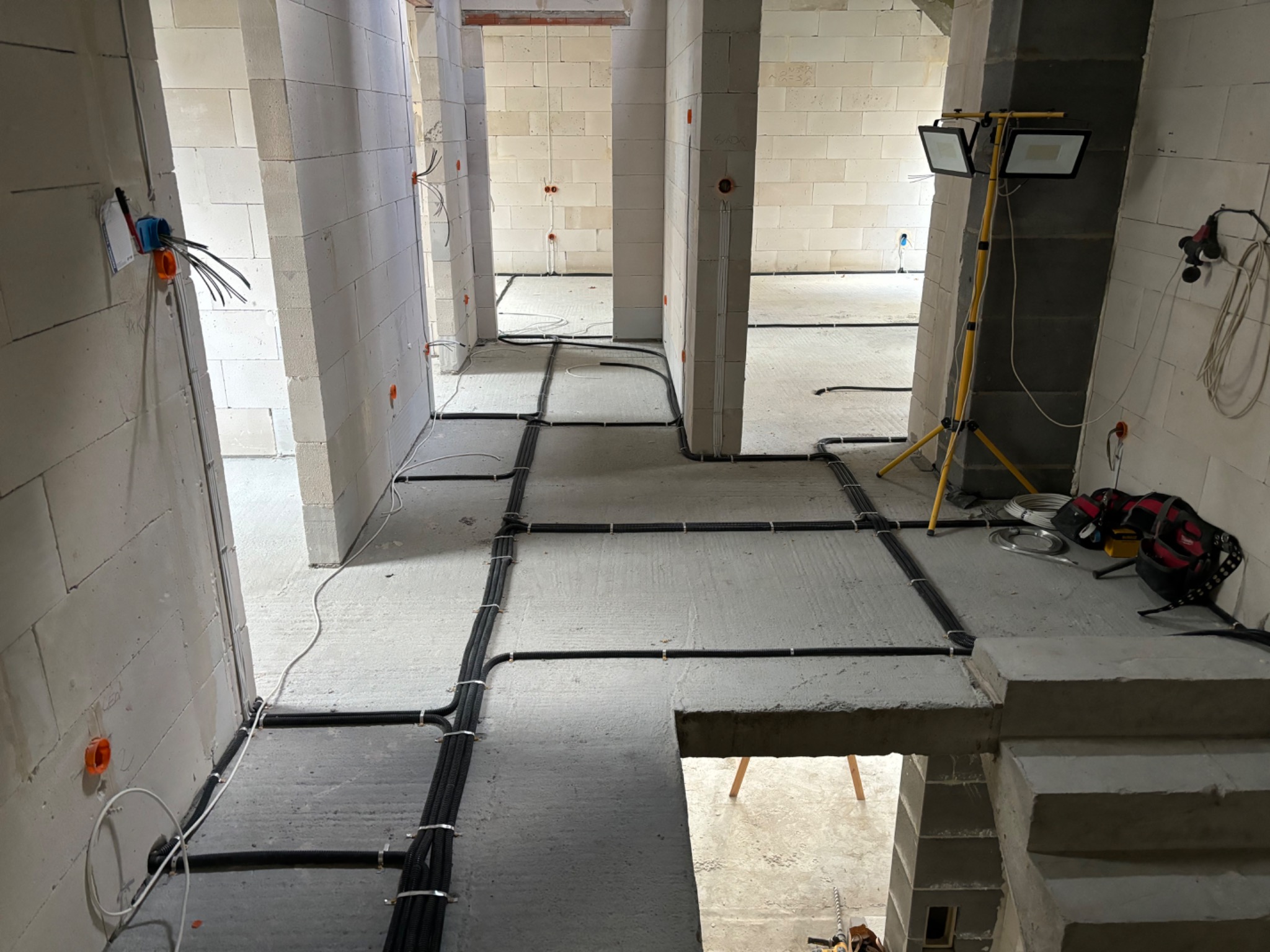 Rozprowadzone czarne kable elektryczne w peszlach, przymocowane do surowego betonowego podłoża w budynku w stanie deweloperskim, widoczne puszki instalacyjne w ścianach z betonu komórkowego...