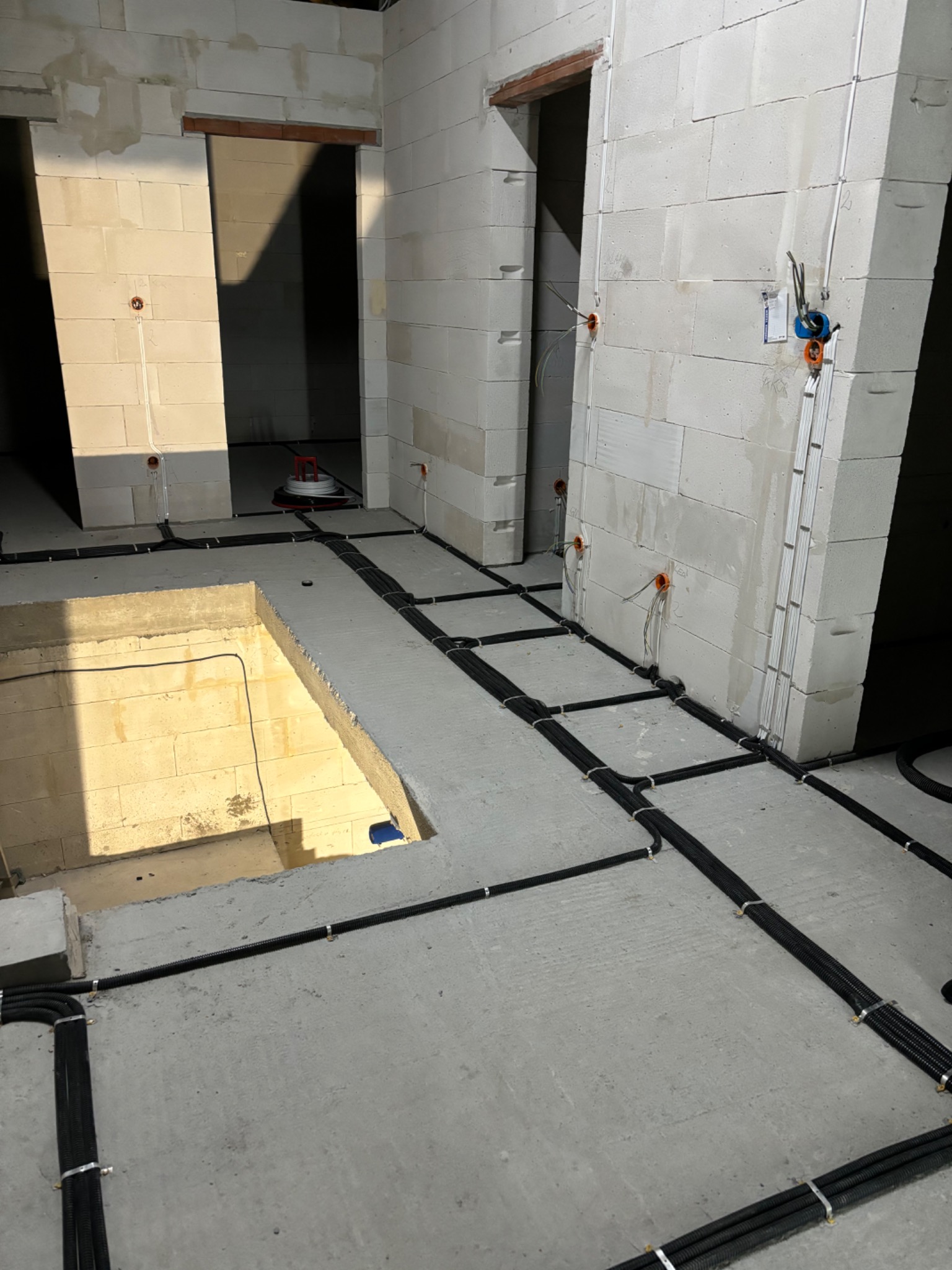 Surowy stan: Czarny peszel z przewodami elektrycznymi ułożony na surowym betonie podłogi w trakcie prac instalacyjnych w budynku z białych bloczków.