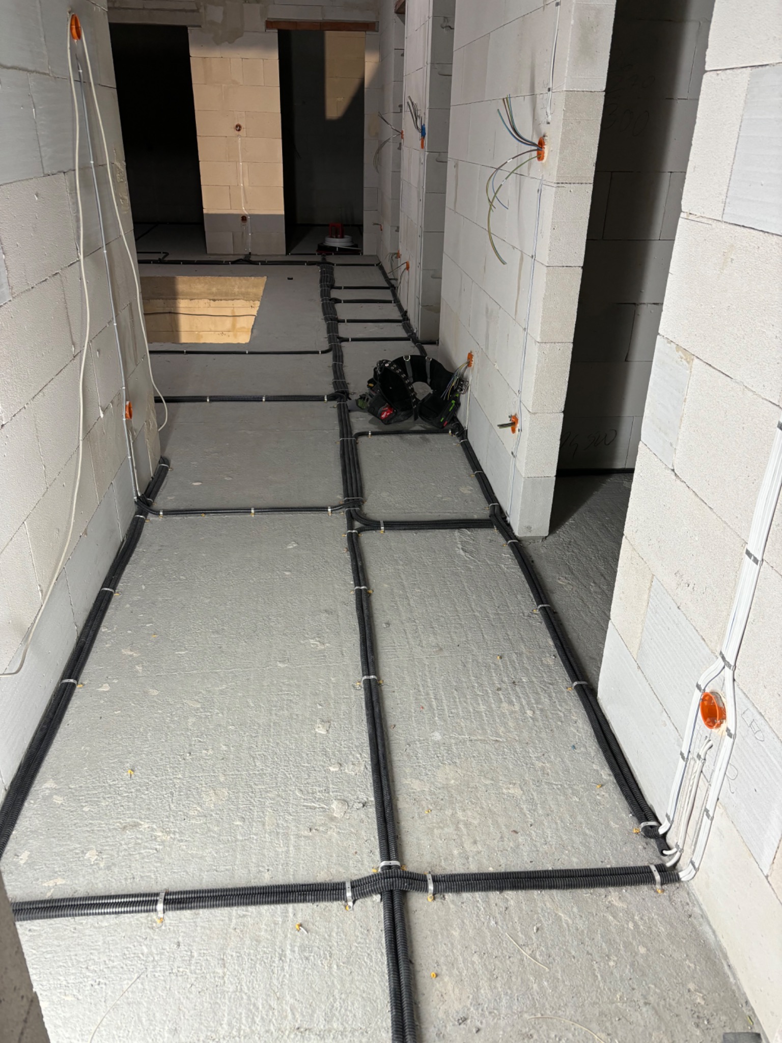 Wnętrze w trakcie budowy z widocznymi czarnymi rurami instalacyjnymi ułożonymi na szarej podłodze, przymocowanymi do podłoża i ścian za pomocą białych uchwytów, z otwartymi puszkami elektrycznymi...