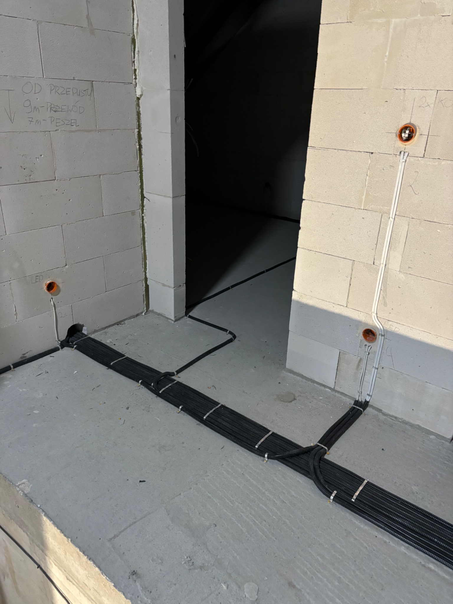 Surowe wnętrze w trakcie prac: kable elektryczne w peszlach ułożone na podłodze, wychodzące ze ściany z widocznymi puszkami instalacyjnymi. Na ścianie widoczne odręczne notatki dotyczące instalacji.