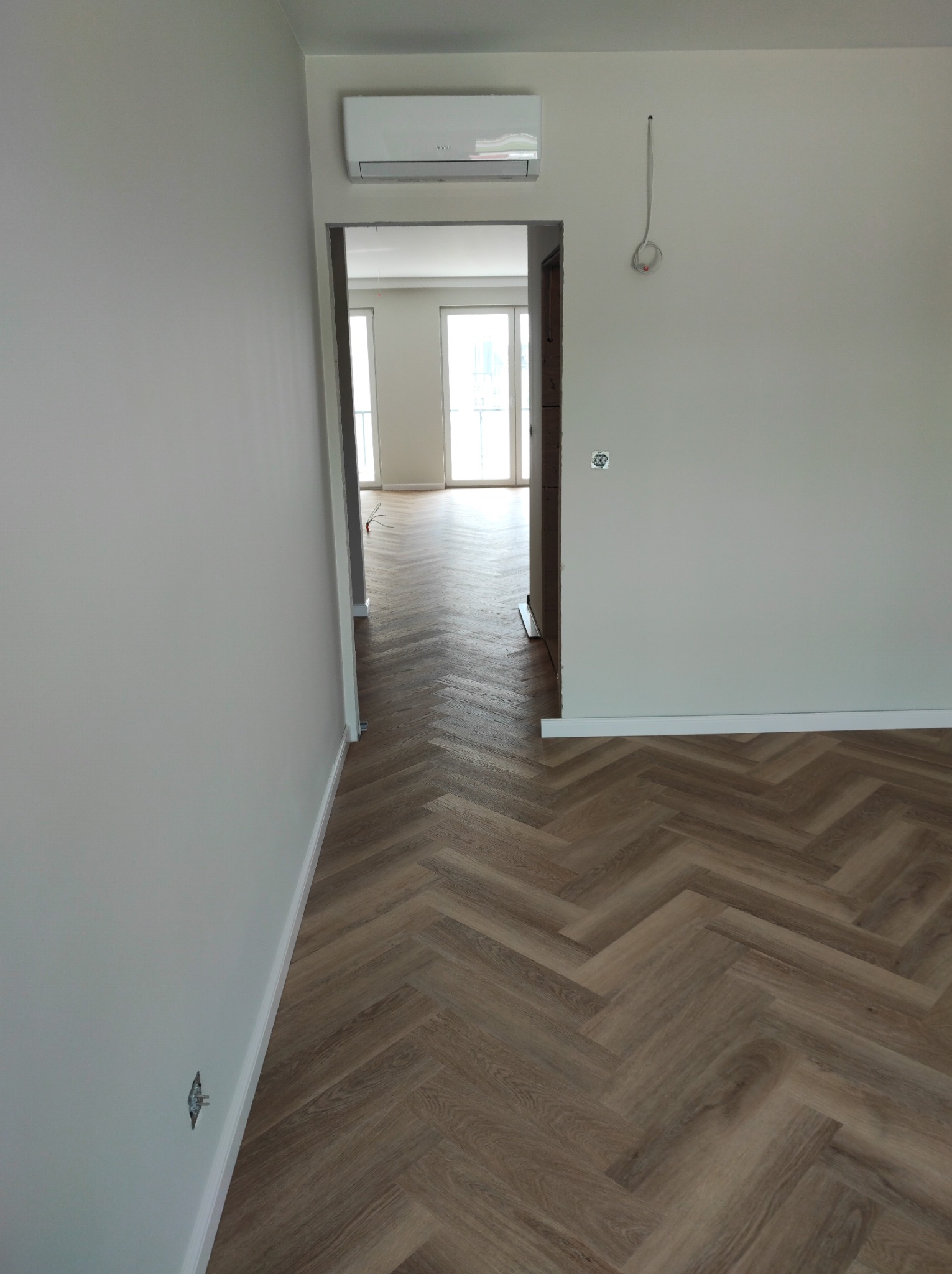 Nowoczesne wnętrze z ułożonym parkietem w jodełkę, widoczne białe ściany, listwy przypodłogowe, klimatyzator i niedokończone okablowanie elektryczne.
