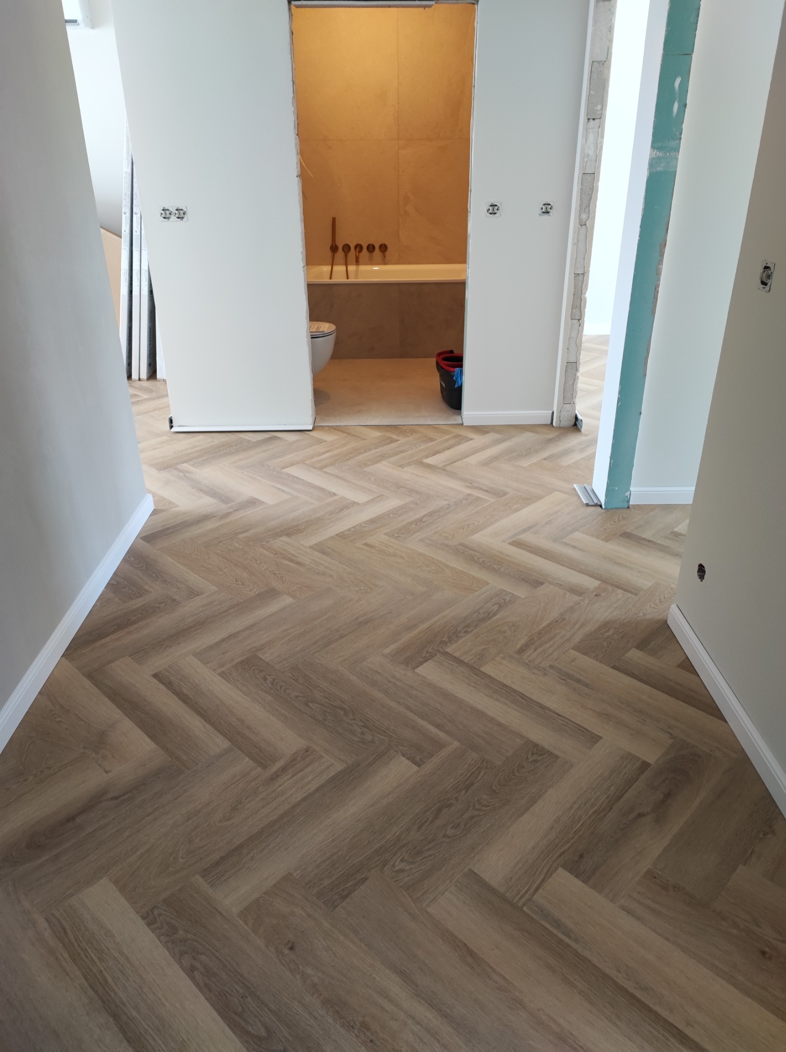 Ułożony parkiet w jodełkę francuską w jasnym odcieniu drewna, widoczne fragmenty łazienki z wanną i toaletą w tle, ściany w trakcie wykończenia.