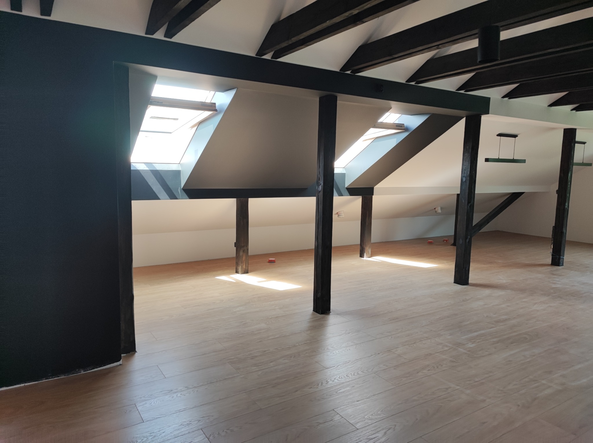 Jasne panele podłogowe w przestronnym pokoju na poddaszu z drewnianymi belkami stropowymi i oknami dachowymi, ściany pomalowane na ciemny kolor, widoczne ślady po pracy.