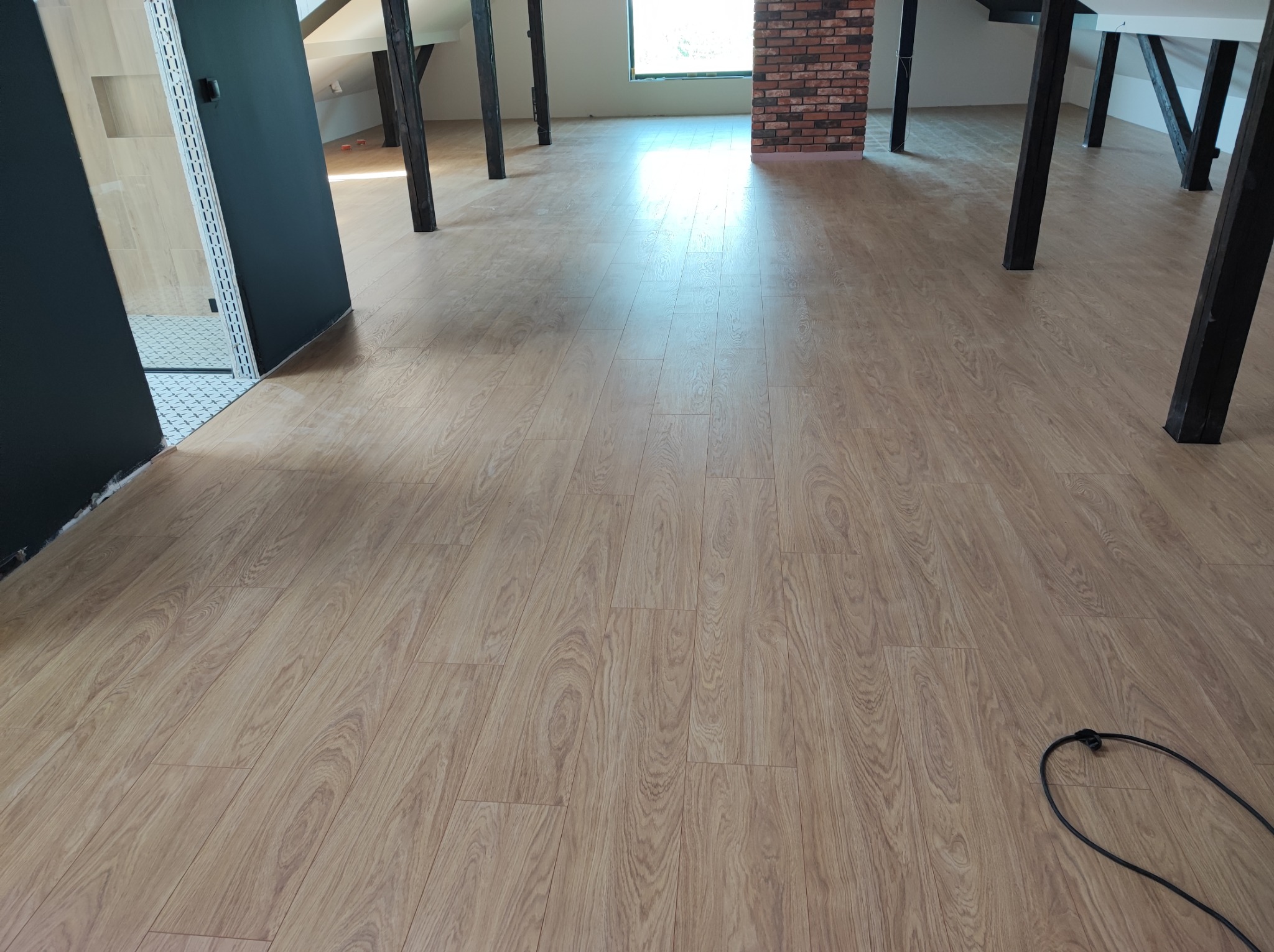 Jasne panele podłogowe ułożone w przestronnym poddaszu z drewnianymi belkami konstrukcyjnymi i wykończonym cegłą kominem, widoczny fragment instalacji elektrycznej przy podłodze.