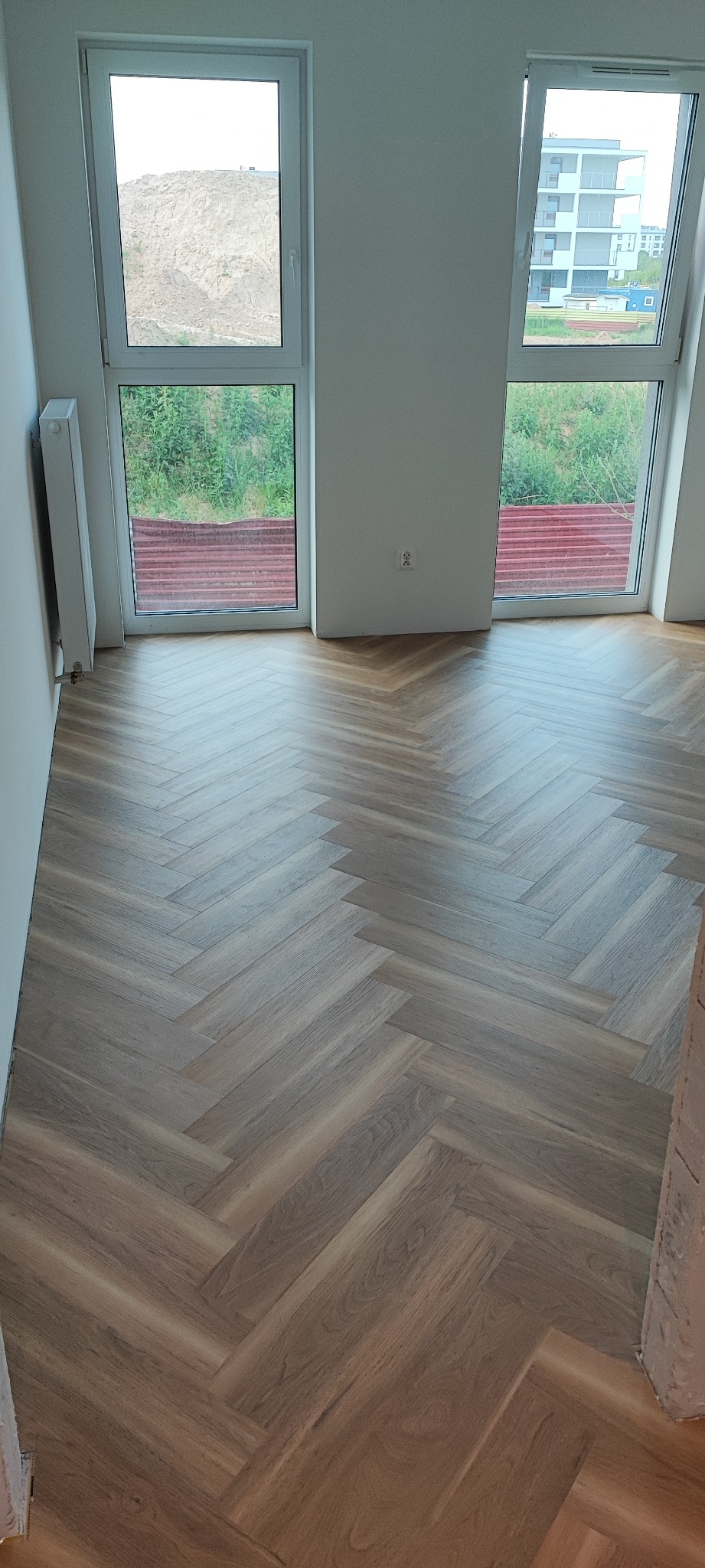 Ułożony parkiet w jodełkę francuską w jasnym, pustym pokoju z dwoma oknami, widok na zewnątrz na wzgórze i budynek.