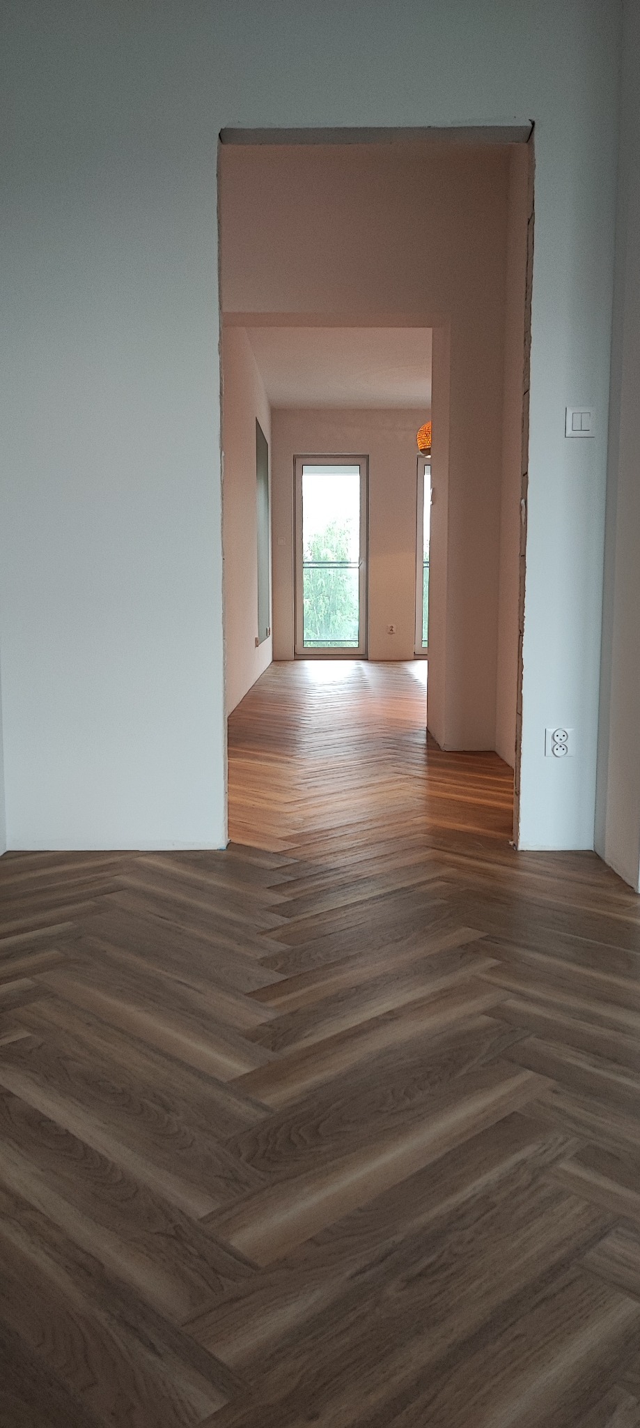 Ułożony parkiet w jodełkę w jasnym pokoju z widokiem na kolejne pomieszczenia przez otwarte przejście, widoczne gniazdka elektryczne na ścianie.