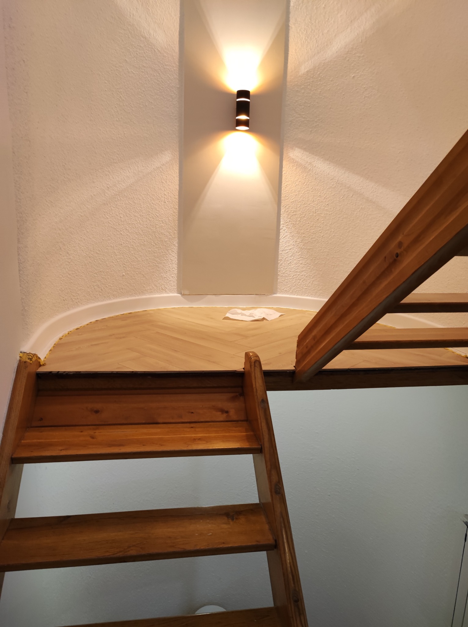 Widok na fragment podłogi z ułożonym parkietem w jodełkę, widziany z góry schodów drewnianych. Ściana z oświetleniem punktowym w tle. Widoczna biała listwa przypodłogowa.