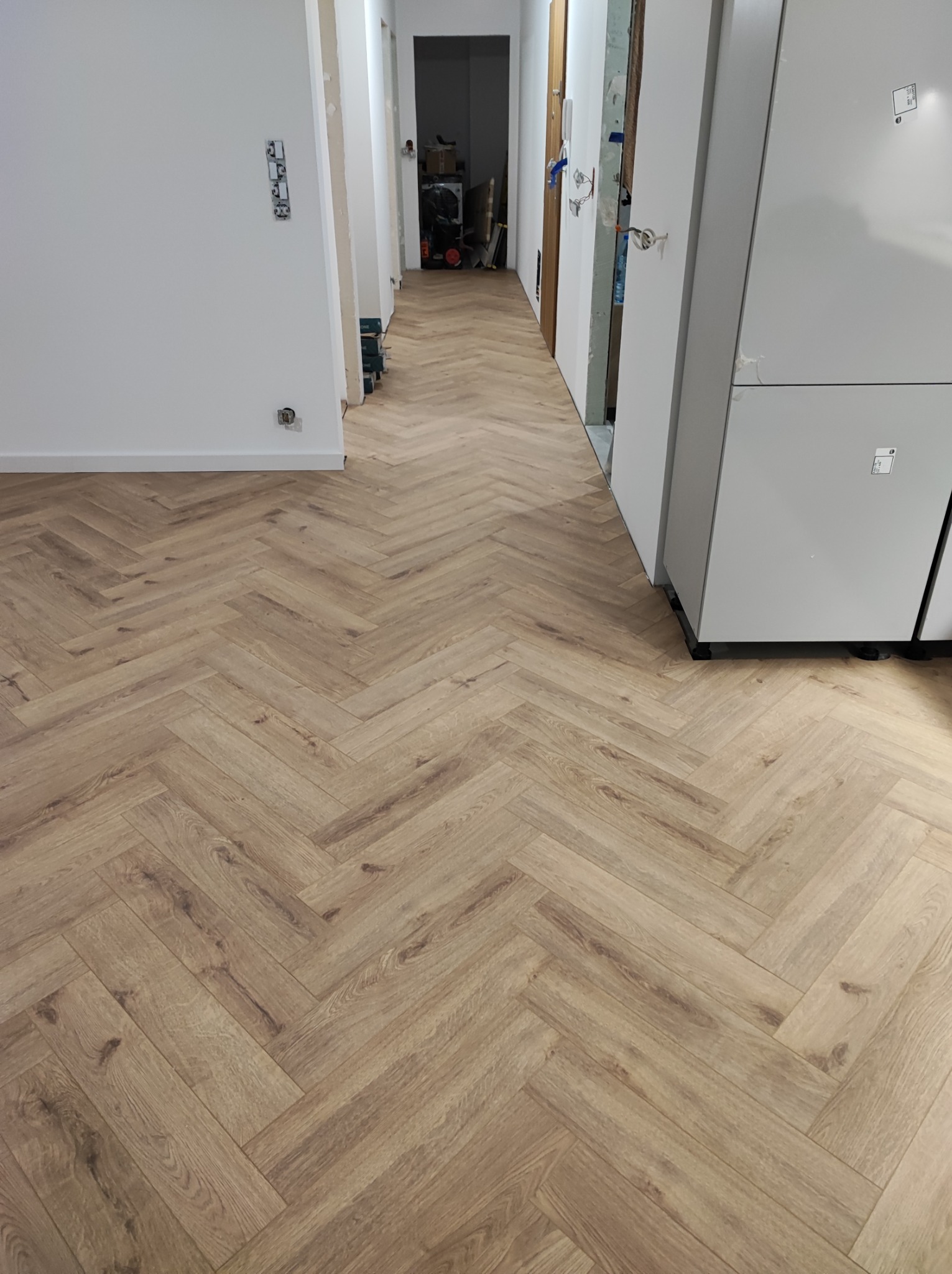 Ułożona podłoga z paneli imitujących drewno w jodełkę francuską, widok z perspektywy wejścia do korytarza, ściany pomalowane na biało, widoczne drzwi w trakcie montażu.