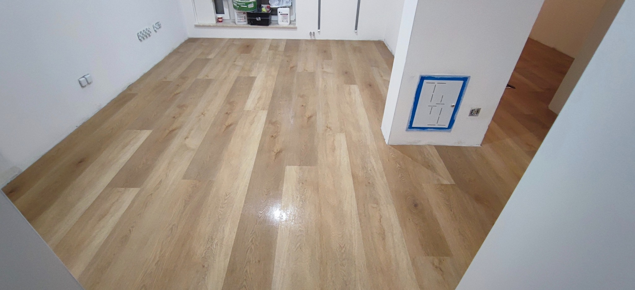 Jasne panele podłogowe imitujące drewno, ułożone w nowym pomieszczeniu z białymi ścianami i widocznymi gniazdkami elektrycznymi oraz skrzynką z niebieskimi obrysami.