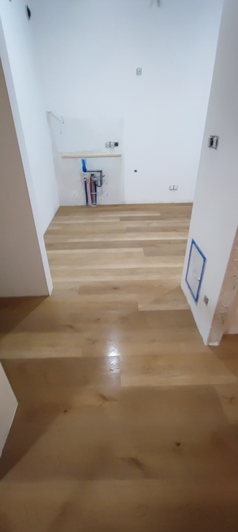 Jasny, świeżo ułożony parkiet w pustym pomieszczeniu z białymi ścianami i widocznymi instalacjami hydraulicznymi w rogu.