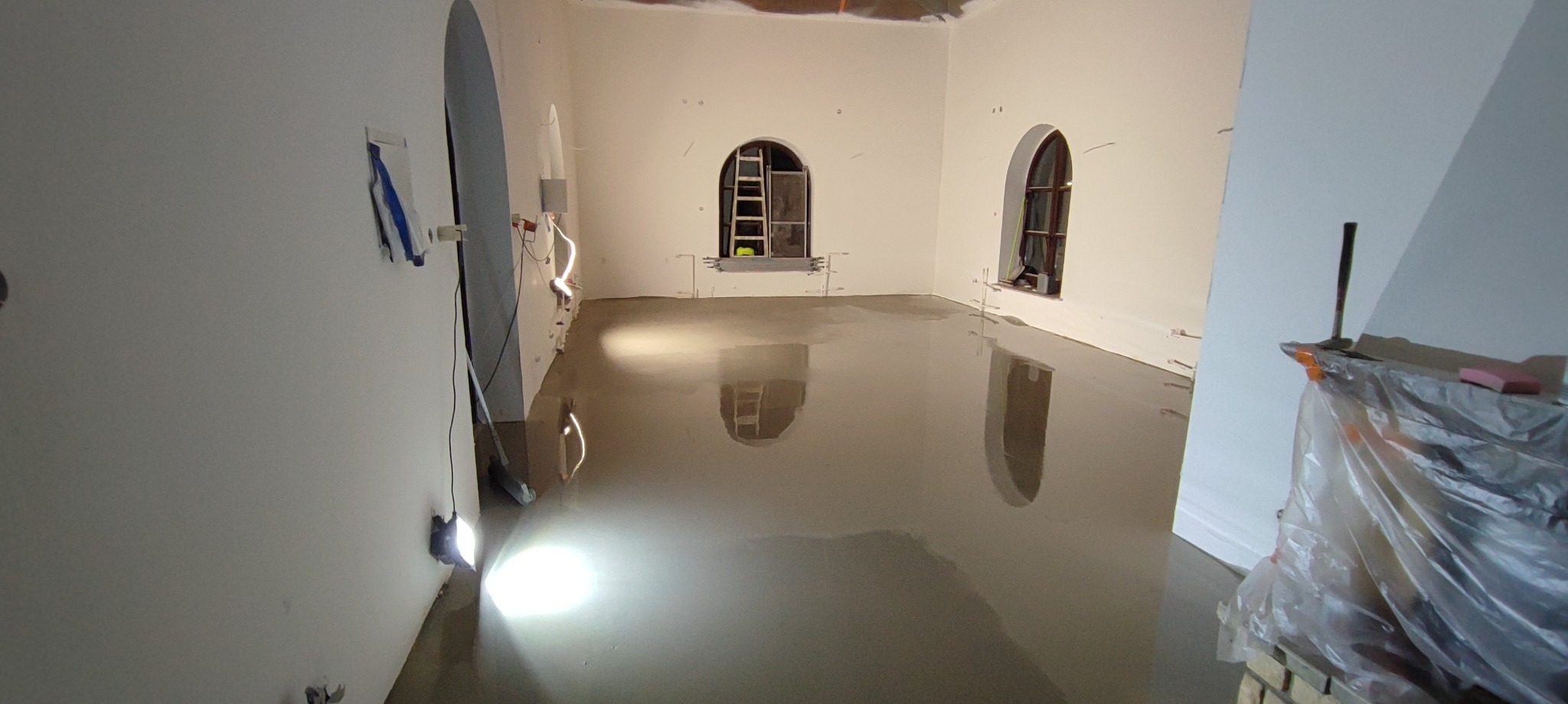 Wylewka samopoziomująca w jasnym pomieszczeniu z oknami łukowymi, widoczny sprzęt budowlany i drabina w tle.