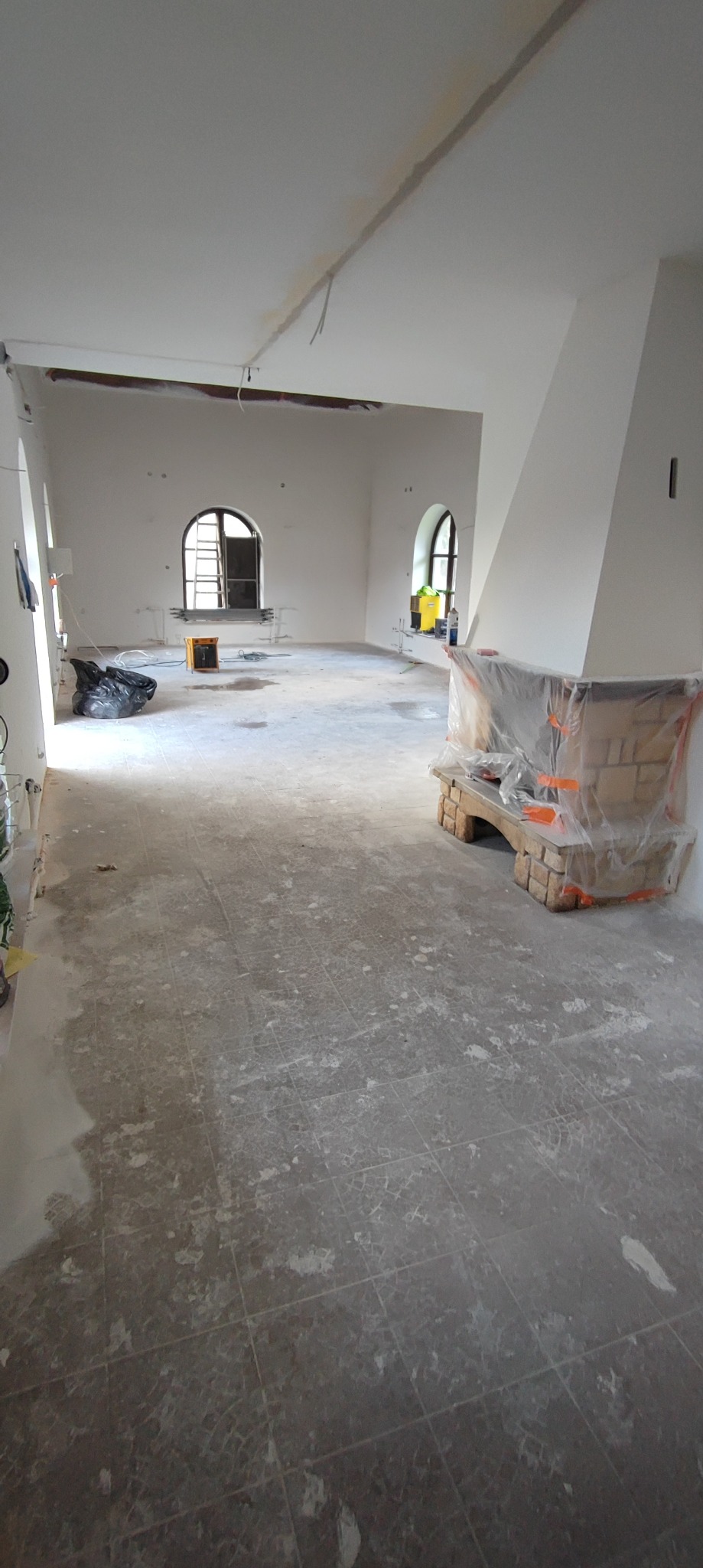 Wnętrze w trakcie remontu: surowe ściany, okna z łukami, zabezpieczony folią kominek z cegły, podłoga z pozostałościami po pracach, widoczny worek z odpadami.