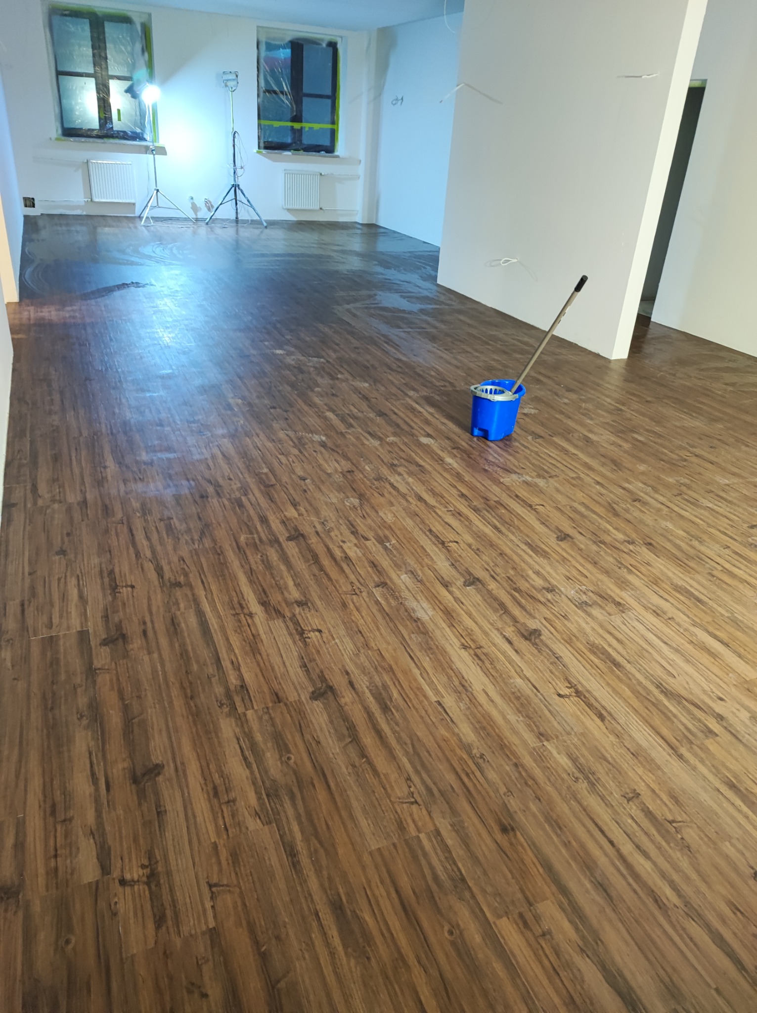 Świeżo ułożona podłoga z paneli imitujących drewno, z widocznym niebieskim wiadrem i mopem, dwa okna zabezpieczone folią malarską, oświetlone lampami budowlanymi.