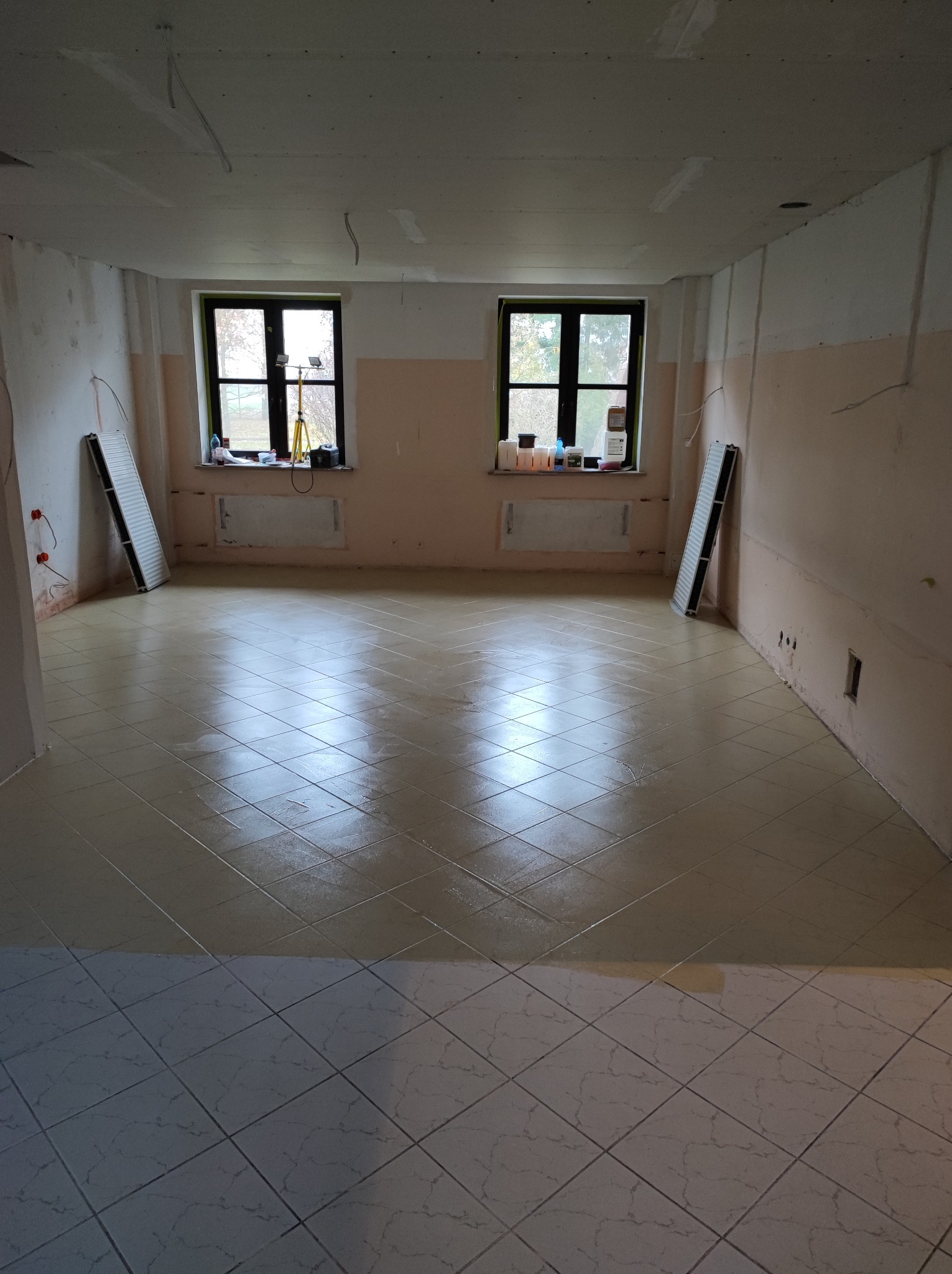 Wnętrze pokoju w trakcie remontu: świeżo położona, błyszcząca podłoga z płytek ułożonych w karo, ściany częściowo pomalowane, widoczne instalacje elektryczne, grzejniki zdjęte, okna z widokiem...