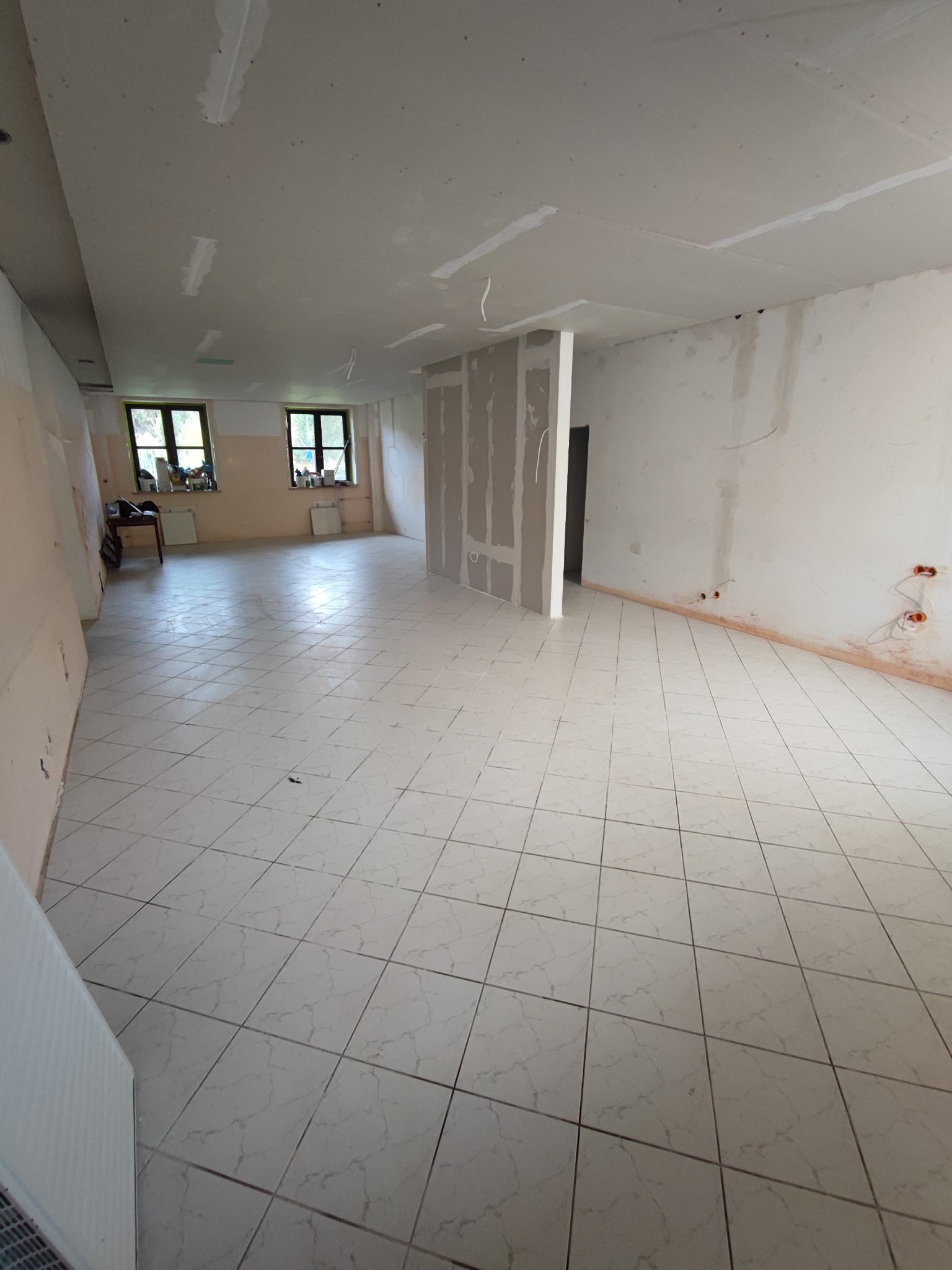 Wnętrze w trakcie remontu: świeżo położone płytki podłogowe, ściany przygotowane do malowania, widoczne instalacje elektryczne, płyty gipsowo-kartonowe na ścianie, okna z parapetami.