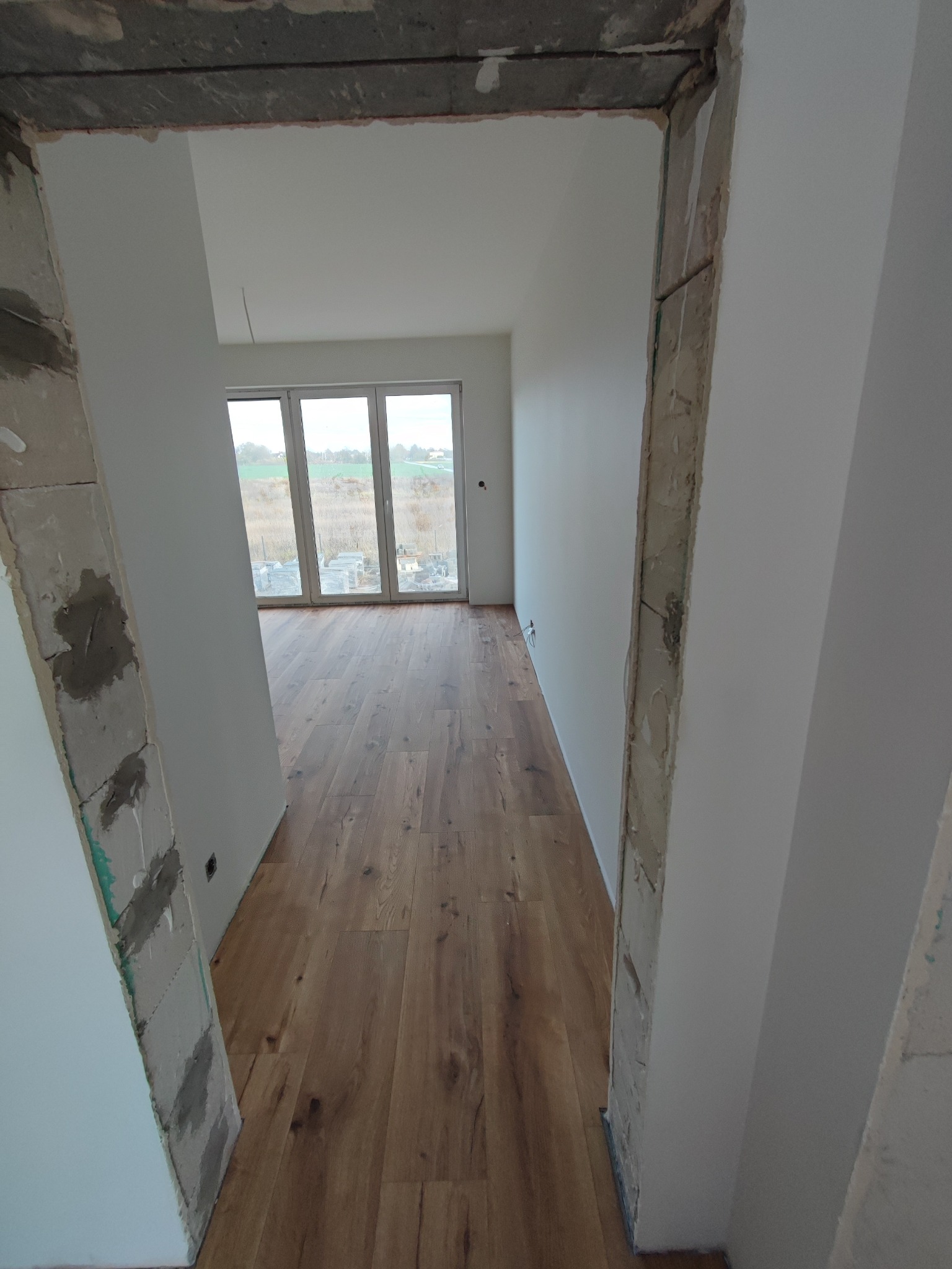 Widok z korytarza na pokój z ułożoną drewnianą podłogą, widoczne surowe wykończenia ścian w przejściu.